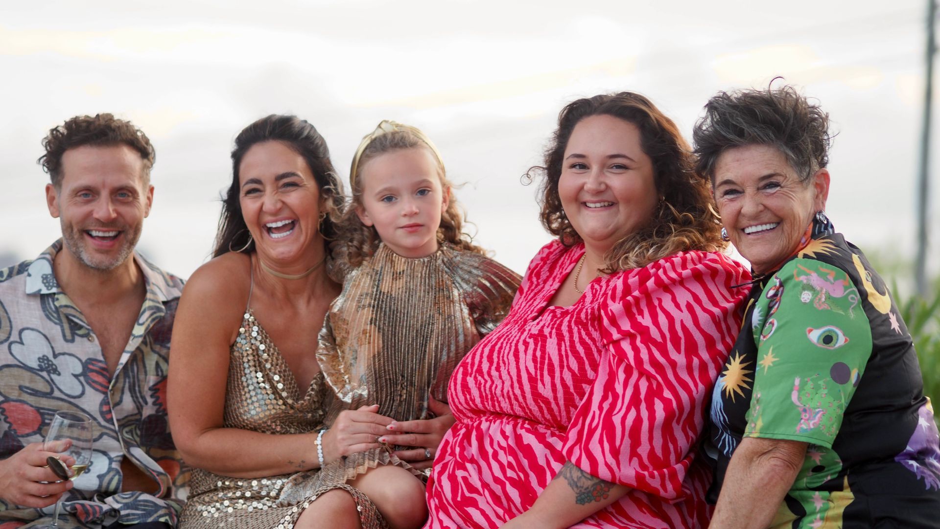 Five people sitting close together outdoors, smiling at the camera, one in a bright pink dress.