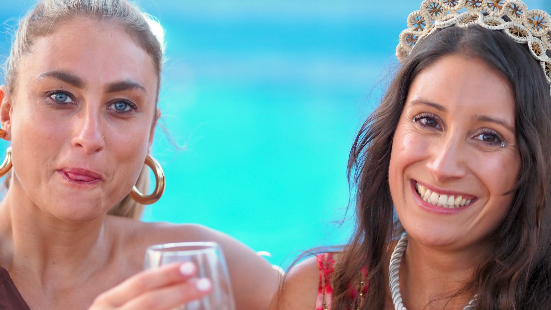 Two smiling women at a beach party, one wearing a tiara and holding a drink, with turquoise water behind them