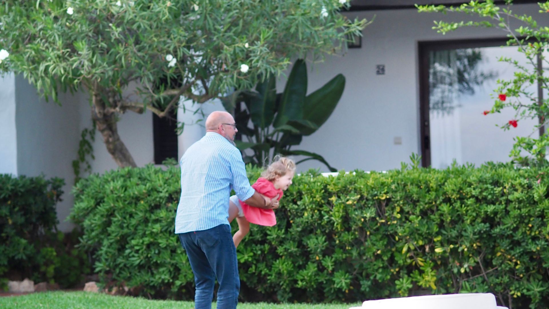 Man helping a child in pink beside a hedge in a suburban yard