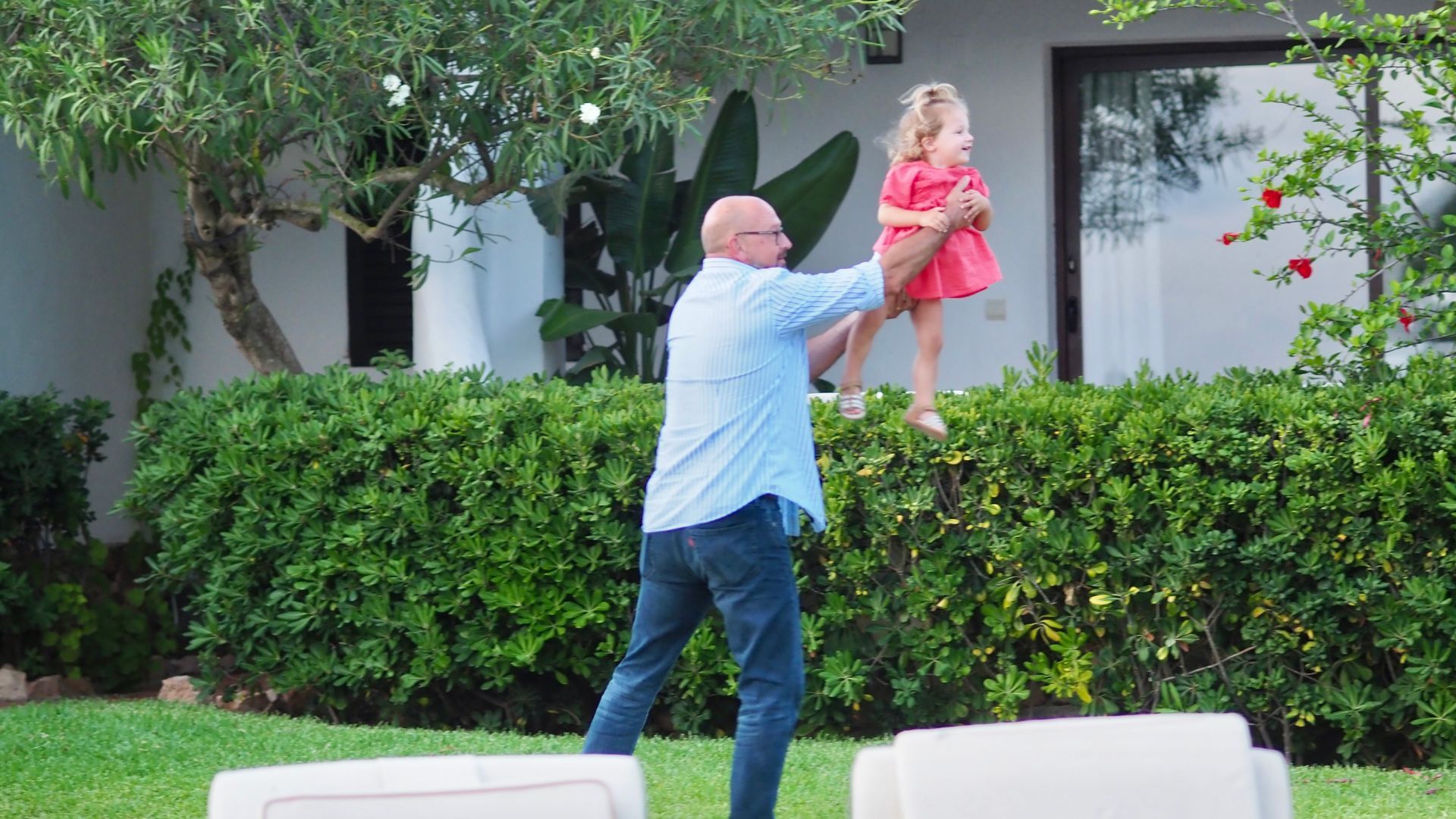 Man in blue shirt lifts a child in a pink dress above a hedge in a garden courtyard.