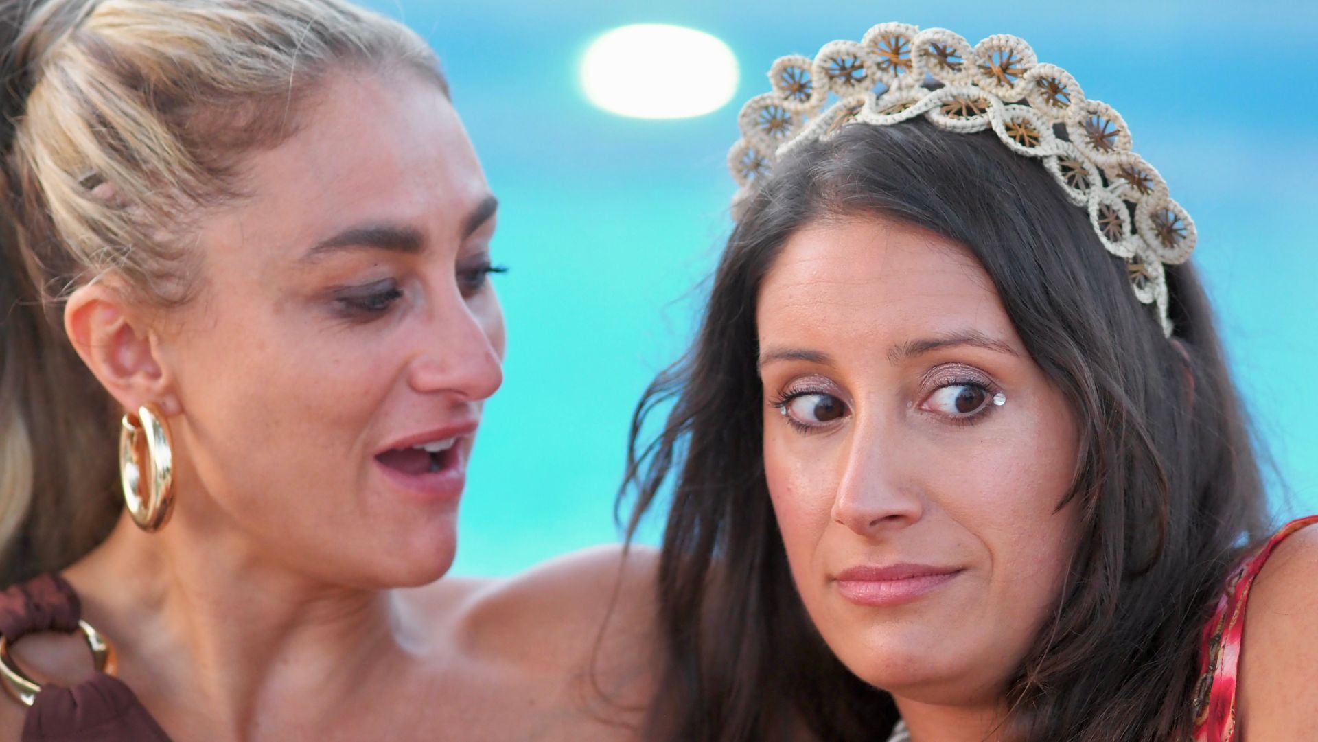 Two women in close-up, one wearing a jeweled tiara, both against a blurred blue background.