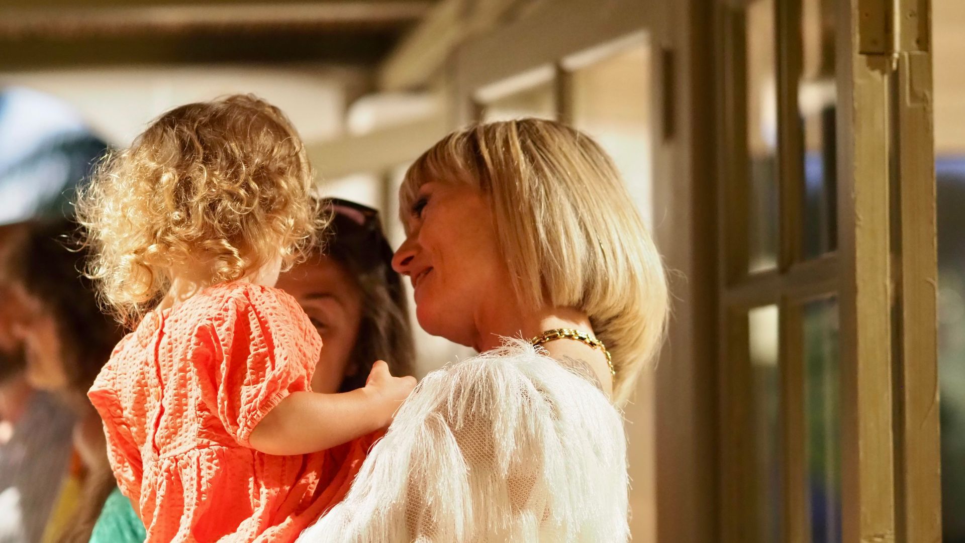 Adult holding a small child in a sunlit hallway, both facing each other near a doorway.