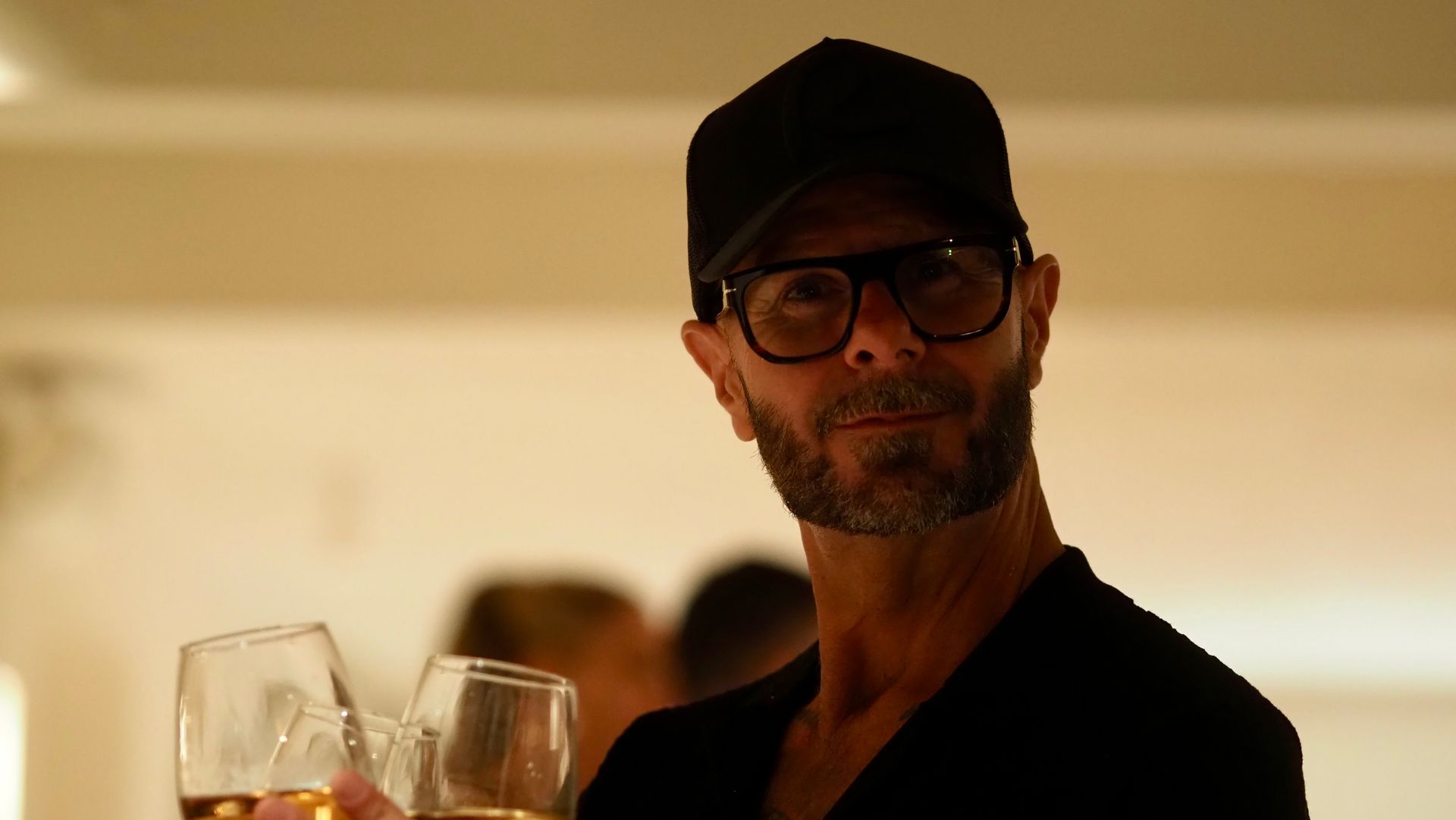 Man in a black cap and glasses holding a drink at a dimly lit event, with people blurred in the background