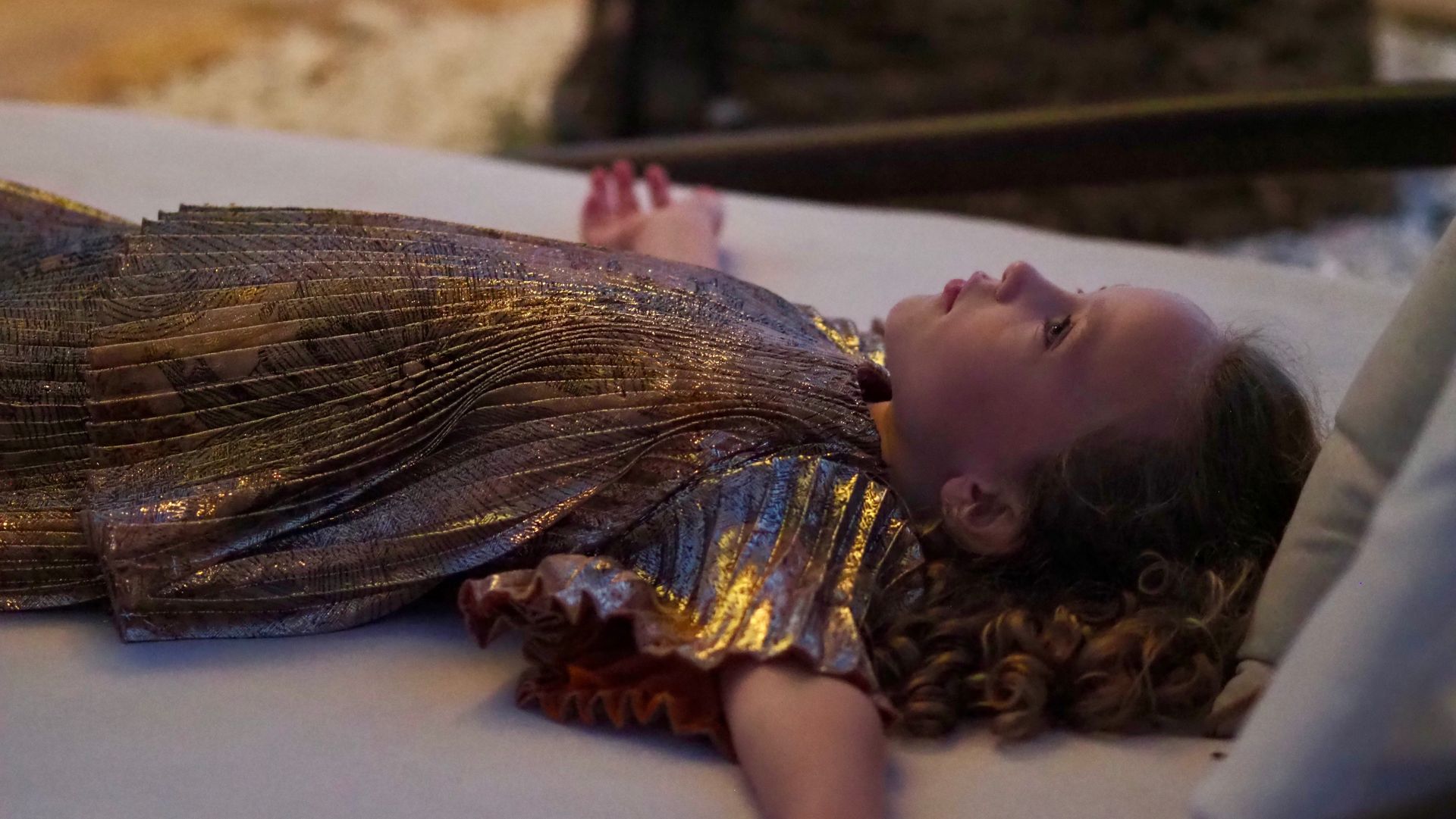 Person in a gold dress lying on a bed, eyes closed, with curly hair and a red flower headband.