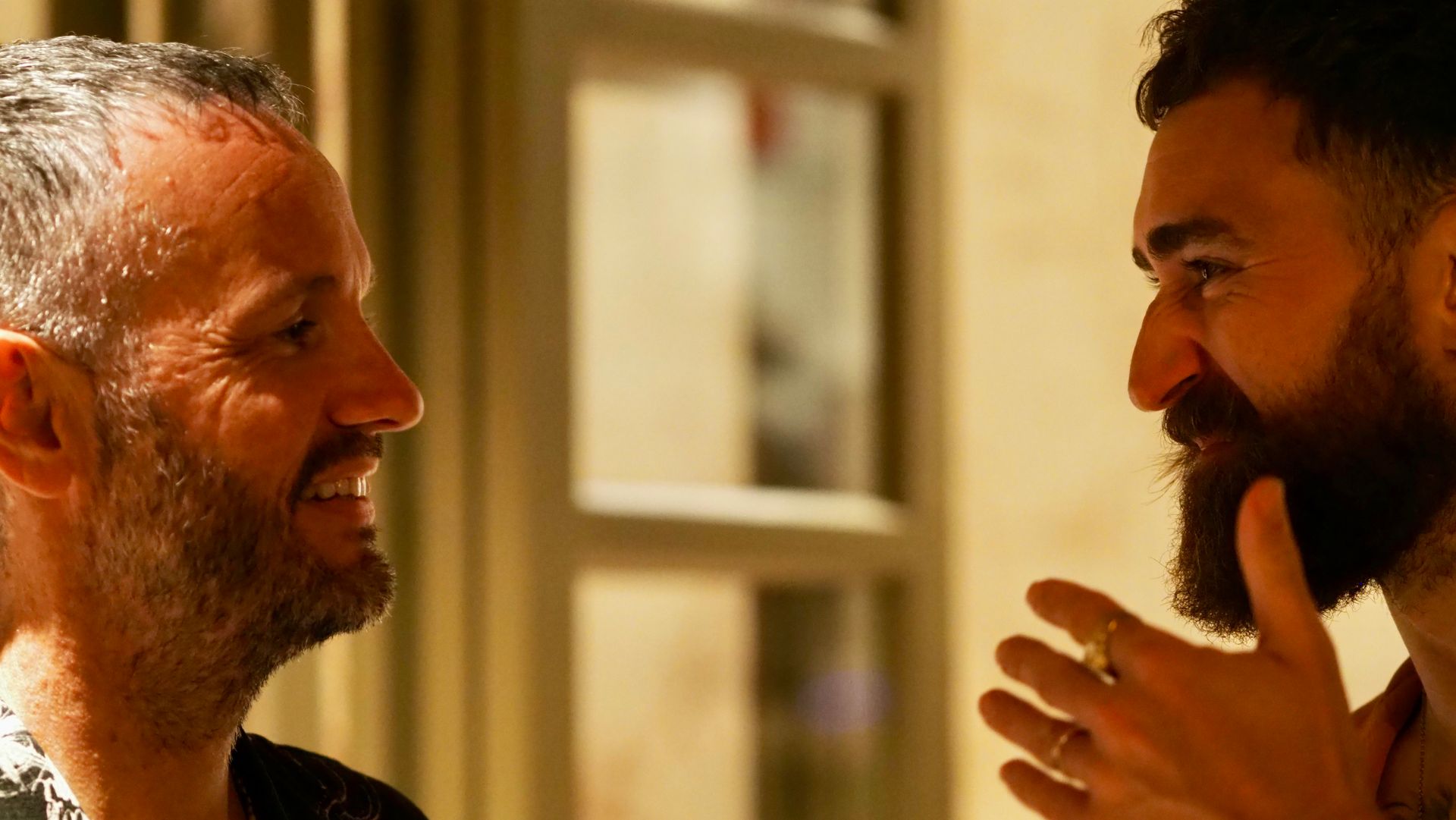 Two men smiling and talking face-to-face indoors in warm light
