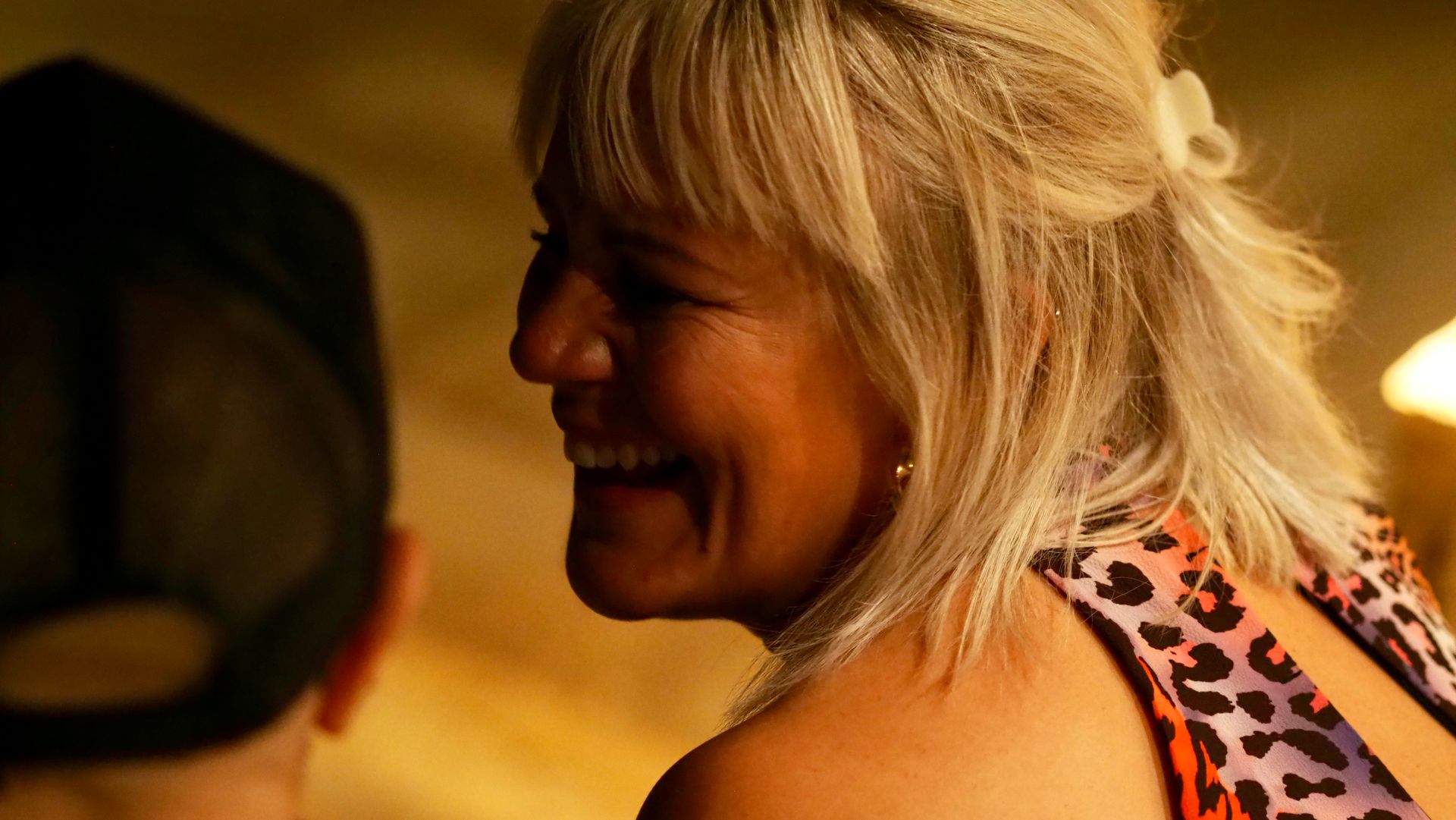 Woman smiling in profile with blonde hair, wearing a leopard-print top in warm indoor lighting