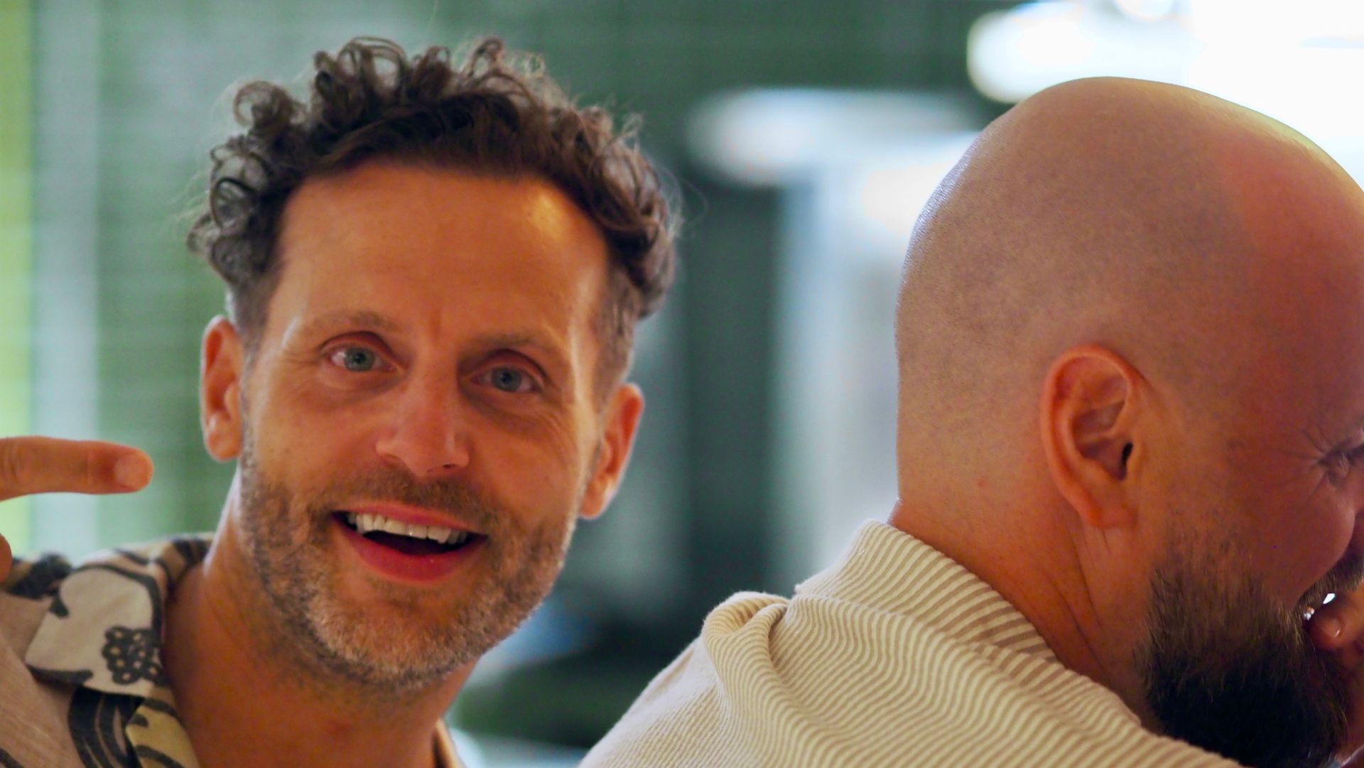 Two men indoors; one smiles at the camera while the other faces away.