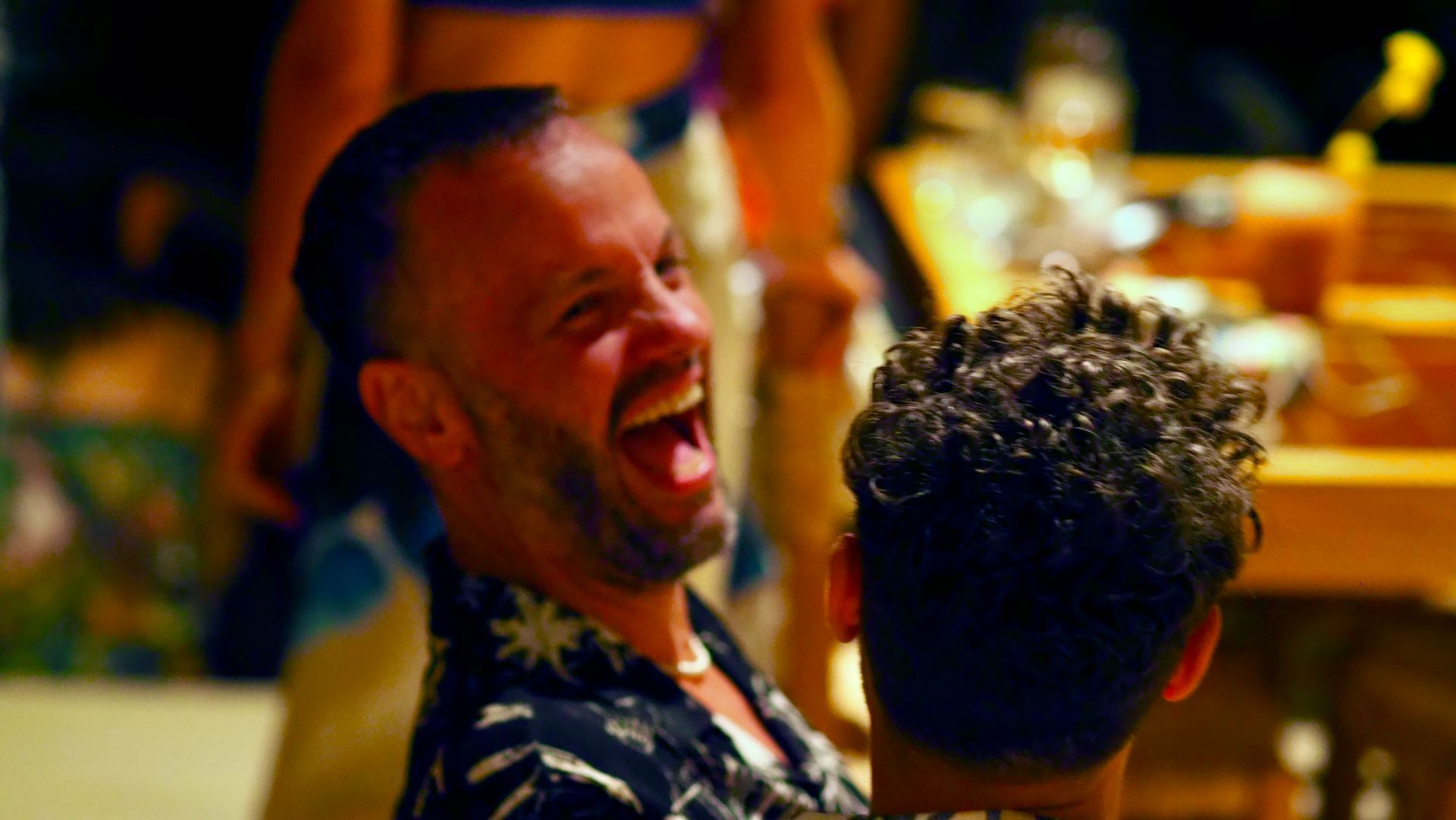 A laughing man in a dark patterned shirt at a warmly lit party, with another person in the foreground.