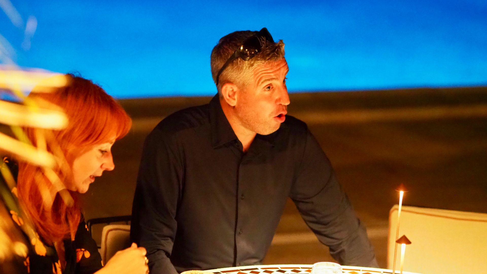 Two people seated at a table, lit by a candle, looking toward a laptop in a warm indoor setting