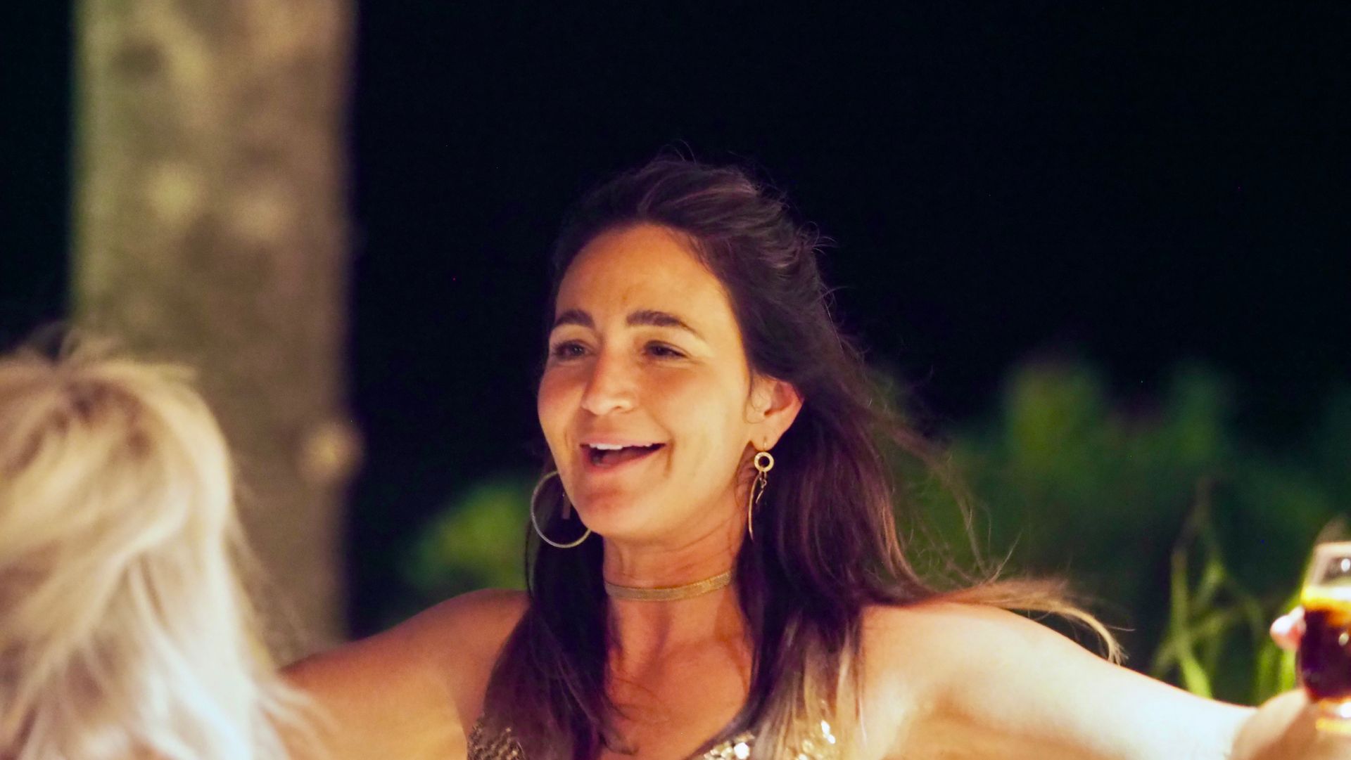 Woman smiling at an outdoor evening gathering, with a blurred blond person beside her and warm lights in the background