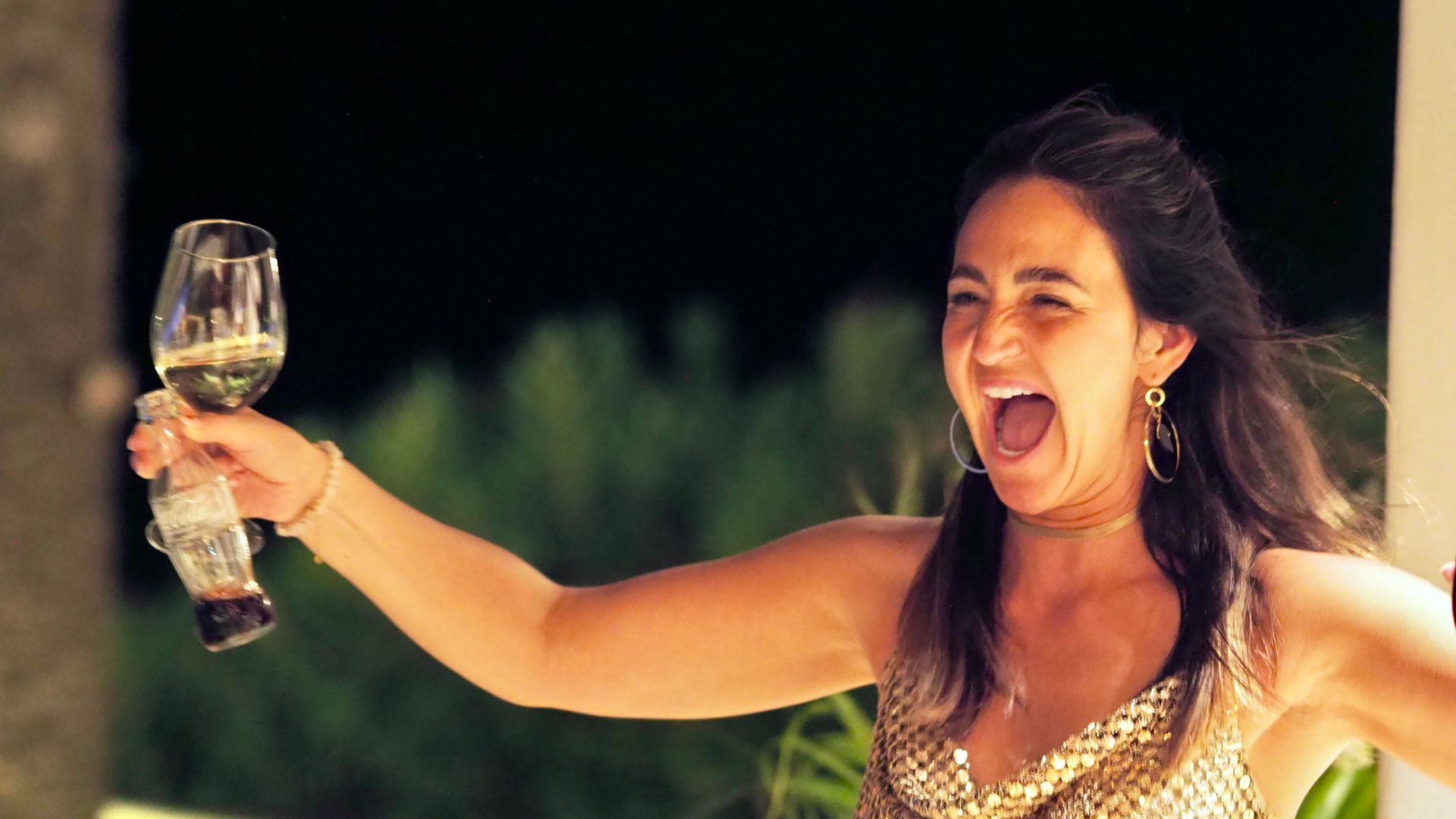 Woman in a gold dress toasting with a glass at an outdoor celebration, smiling and laughing at night