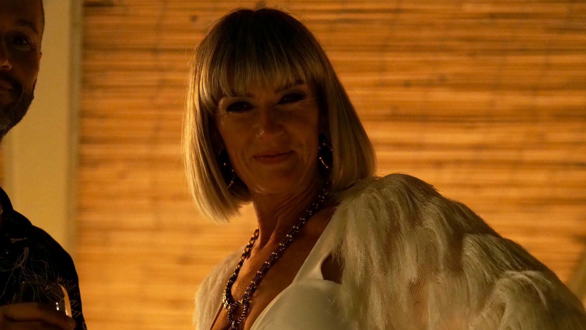 Woman in a white feathered wrap standing in warm amber light, with a curtain backdrop.