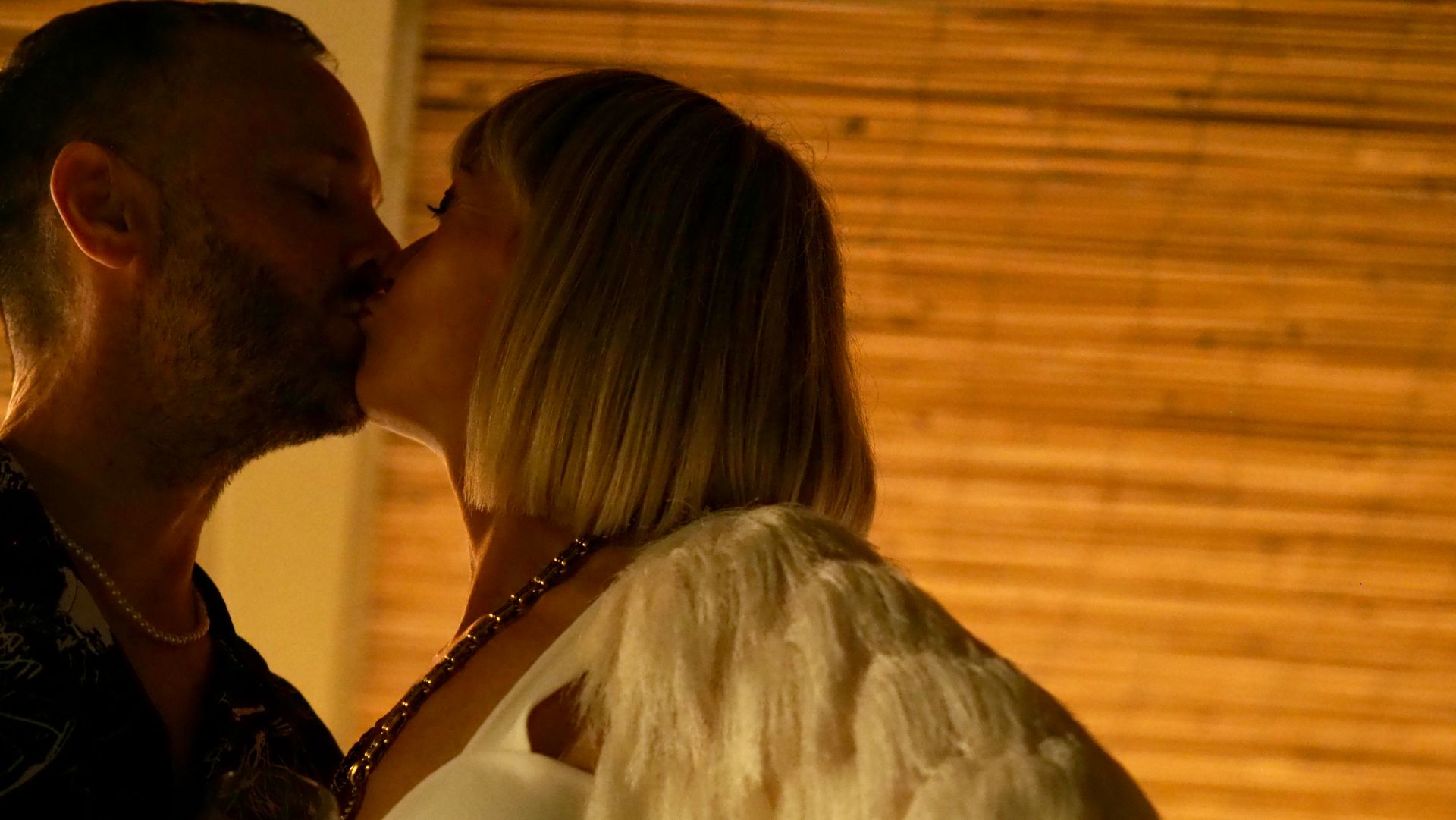 Two people kissing in warm, dim indoor light, with a bamboo wall behind them.