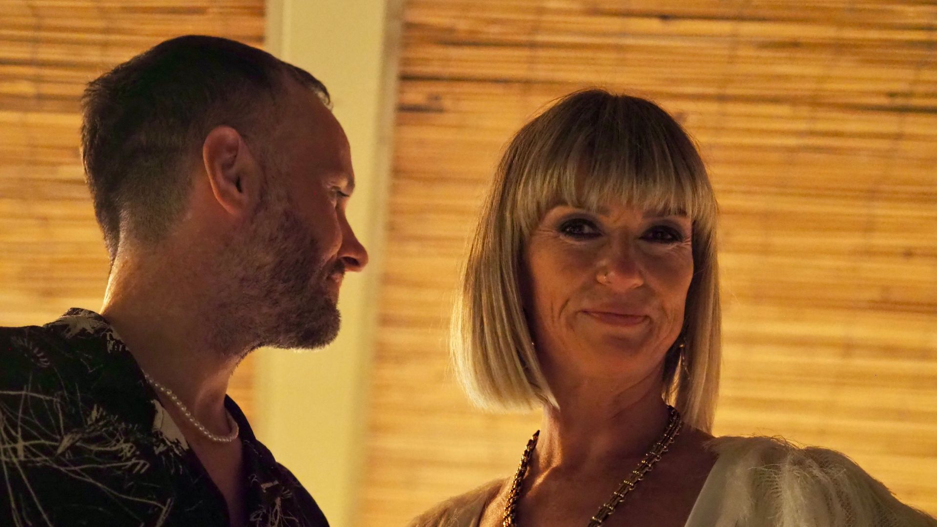 Two people in warm indoor lighting, one facing the other and smiling slightly.