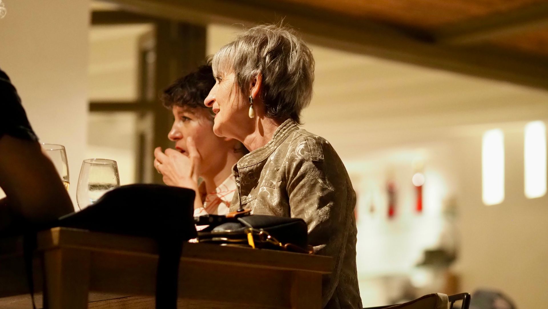 Two people seated at a warmly lit table in a restaurant, talking over drinks and dishes.