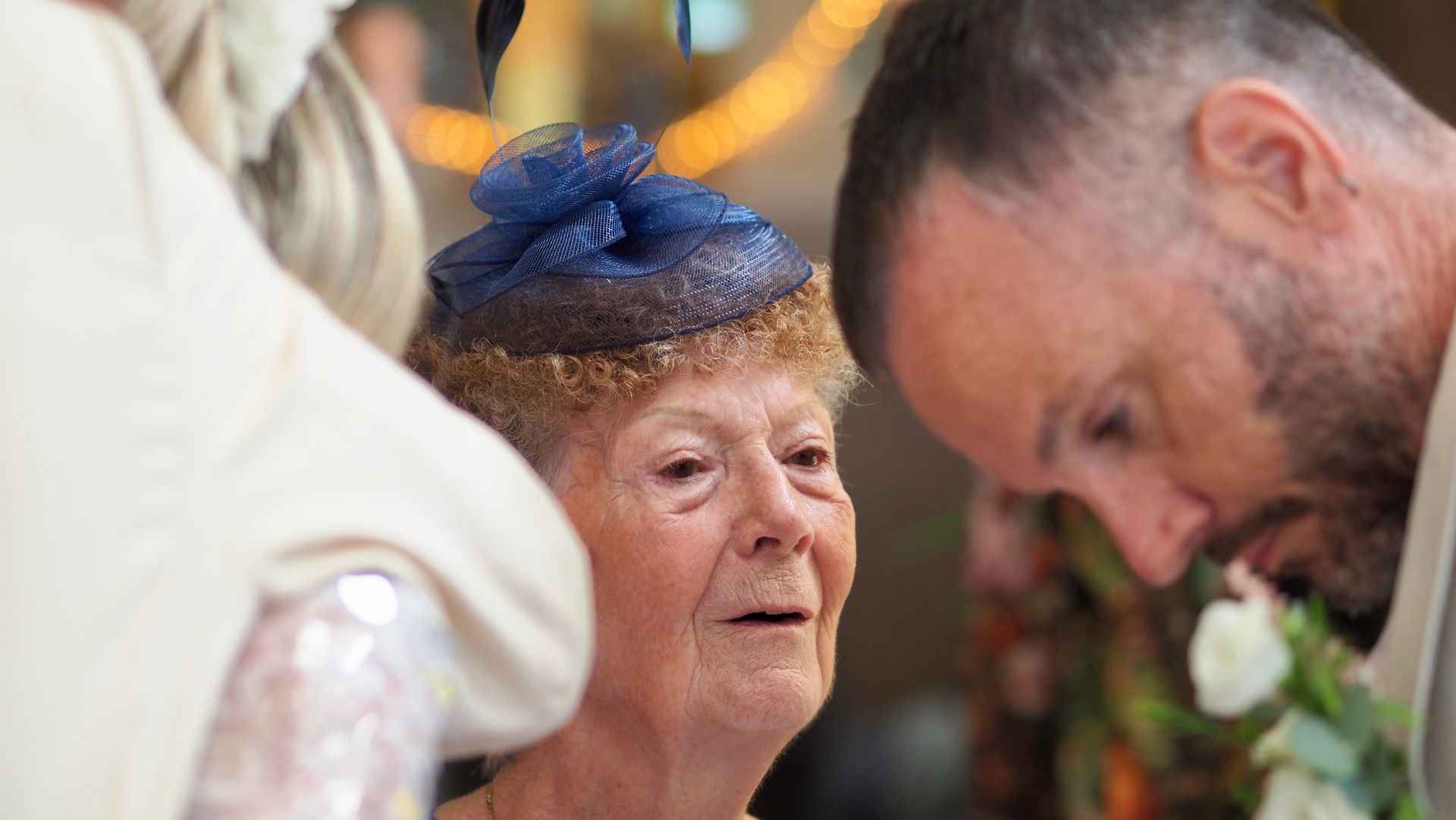 Woman in blue fascinator smiling at a formal event, with two people leaning in to speak nearby