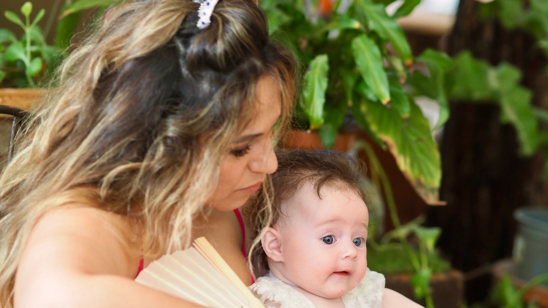 Woman holding a baby outdoors, both looking to the side amid green plants.