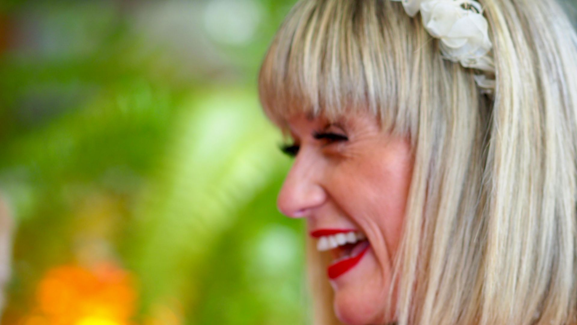 Profile of a smiling blonde woman with red lipstick and a white floral headpiece against a blurred green background