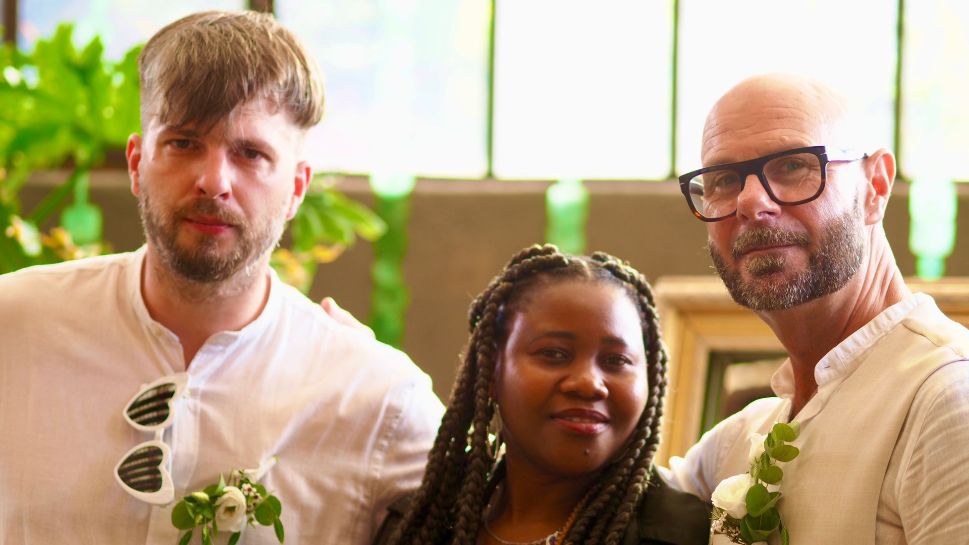 Three people posing indoors with drinks, wearing white shirts and flower accents, in front of a bright window.