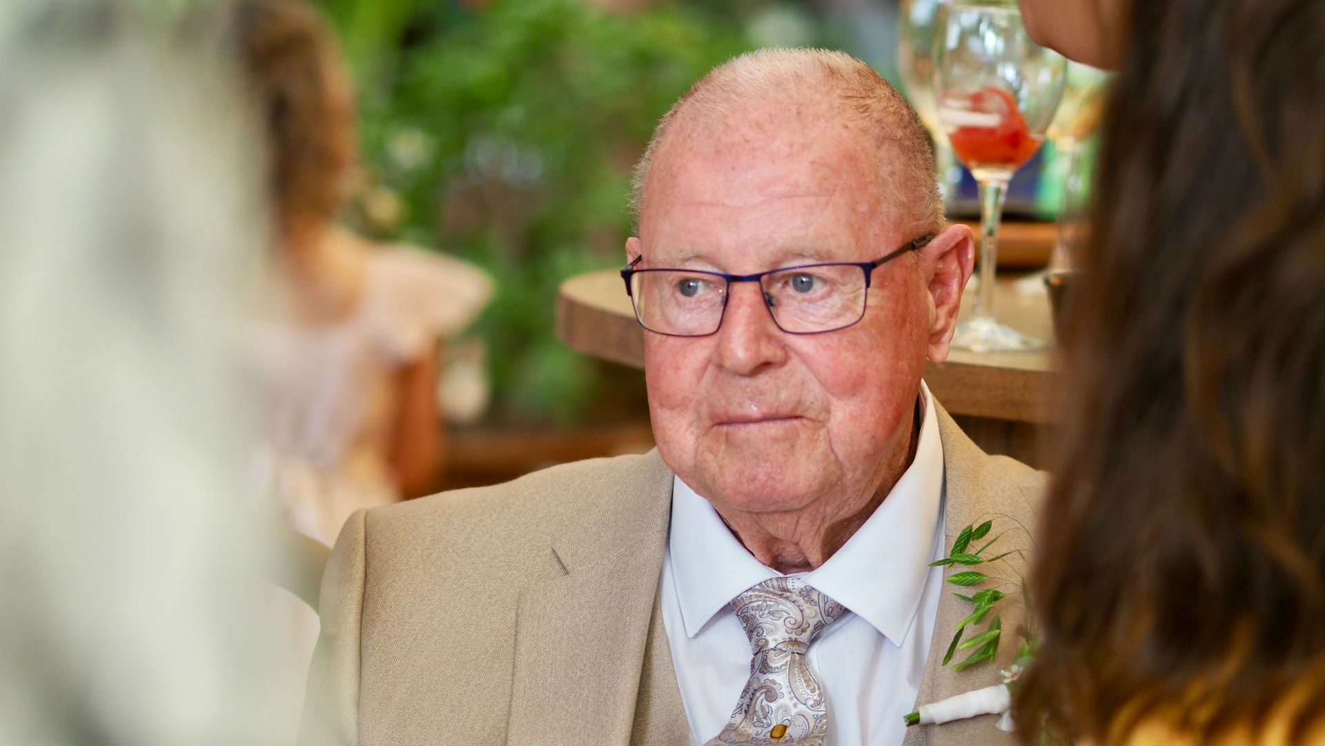 Man in beige suit and glasses seated at a banquet table, listening attentively in a softly lit room
