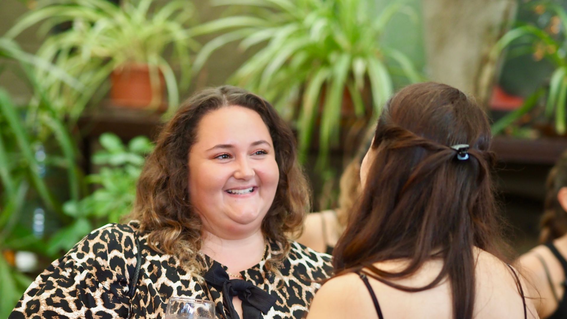 Two women chatting indoors, one smiling, with green plants in the background