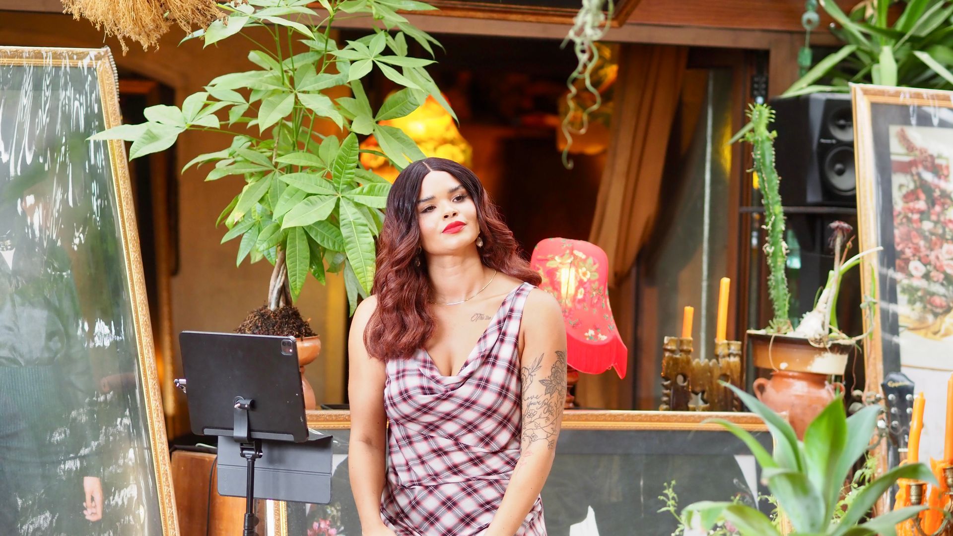Person standing outdoors in a patterned dress near plants and a café counter, looking off to the side.