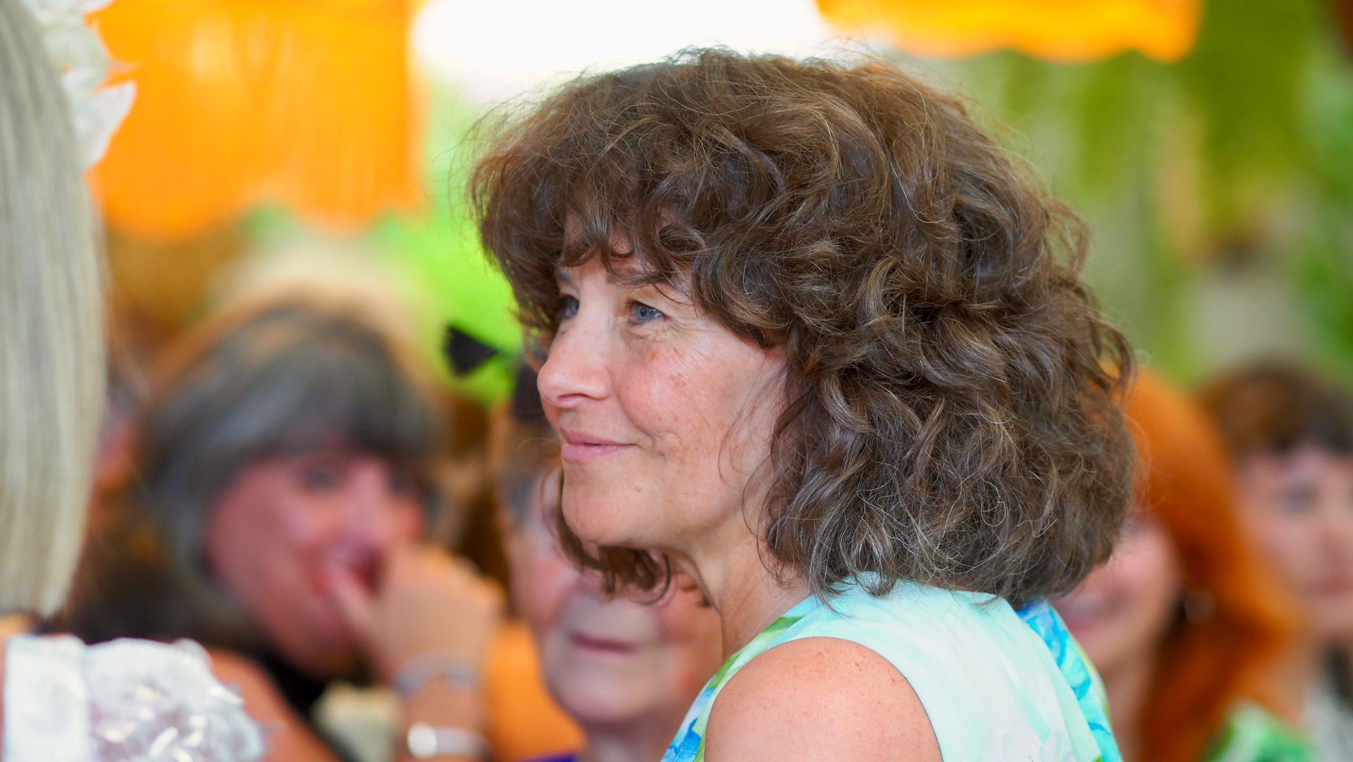Woman with curly brown hair in a turquoise top, smiling in a lively indoor gathering