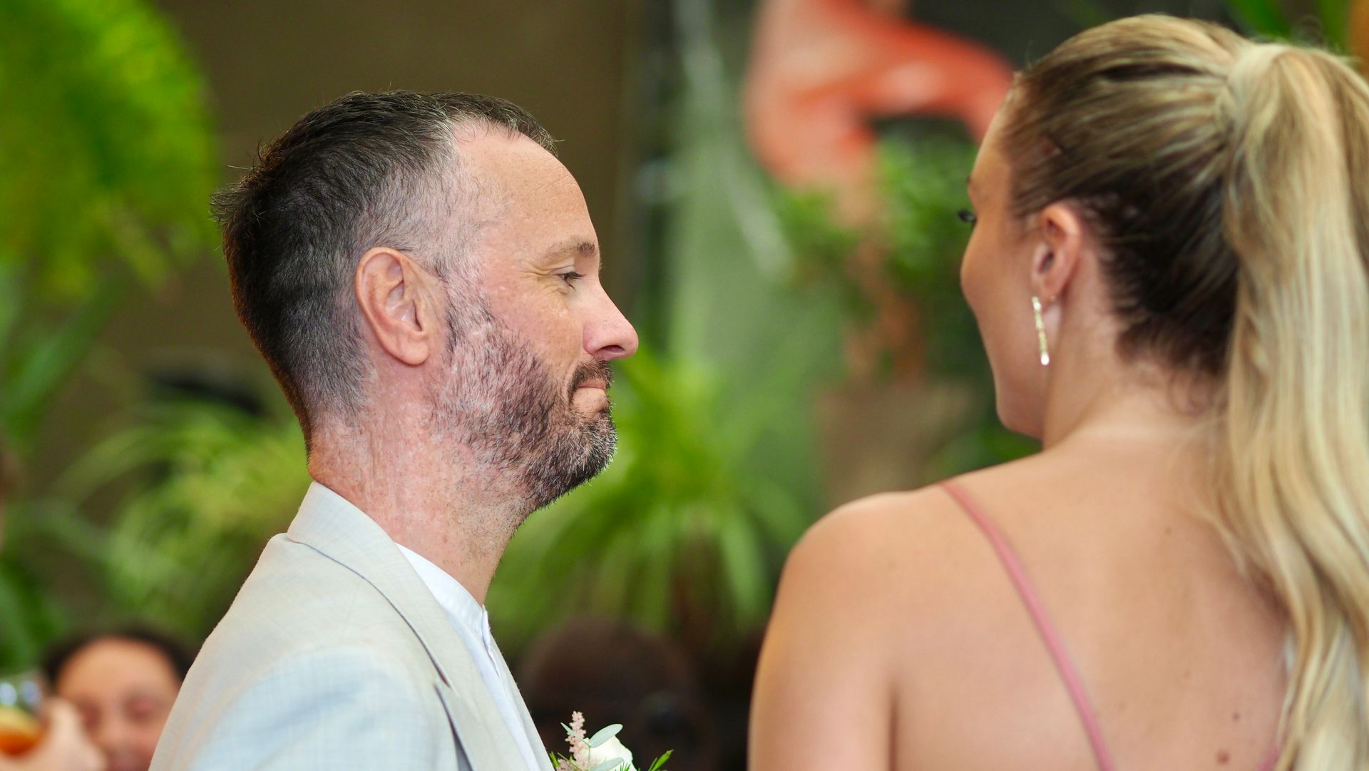 Two people facing each other at a wedding, with greenery and floral decor in the background.