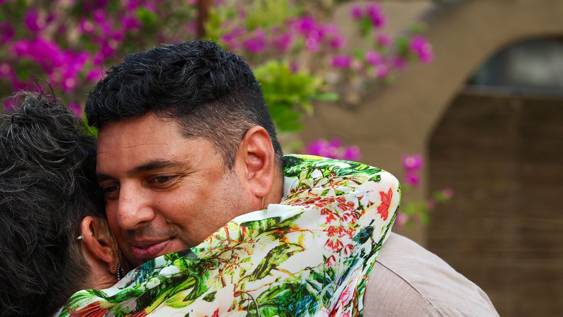 Two people hugging outdoors, with pink flowers in the background.