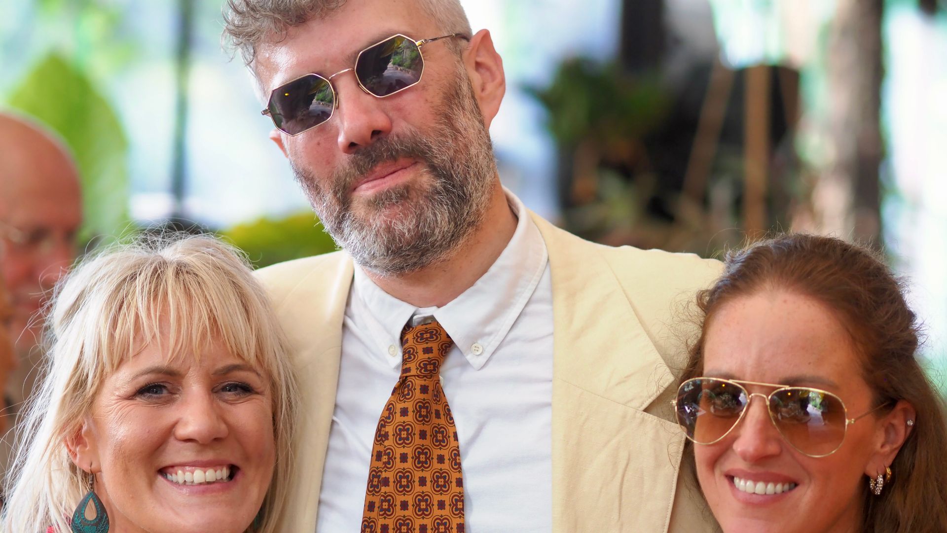 Three people smiling outdoors, with the man in the center wearing sunglasses and a patterned tie.