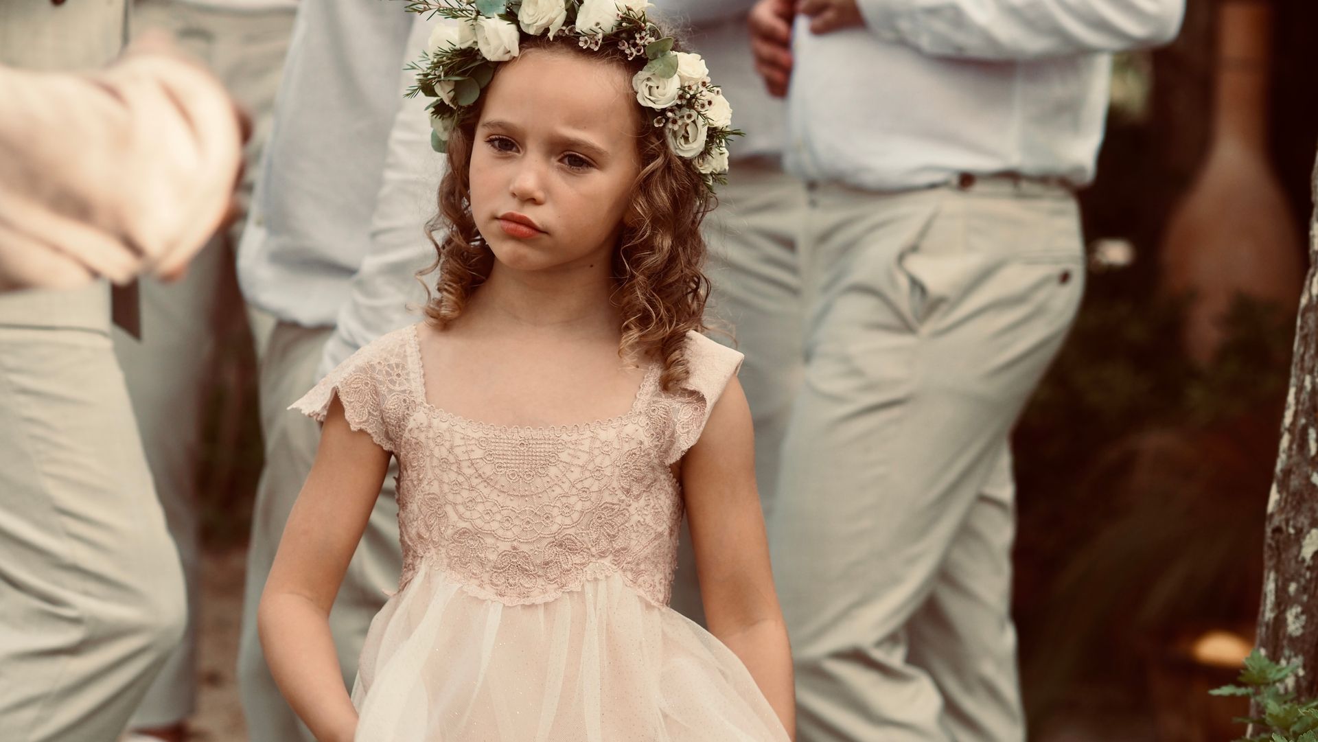 Young girl in a cream dress and flower crown standing outdoors with adults in the background