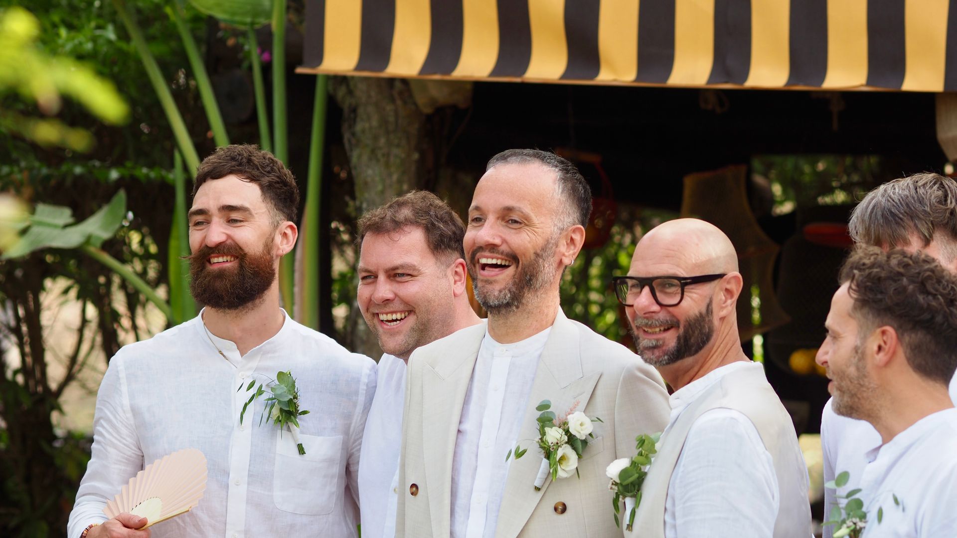 Five smiling men in white suits at an outdoor event, standing under a black-and-yellow awning.