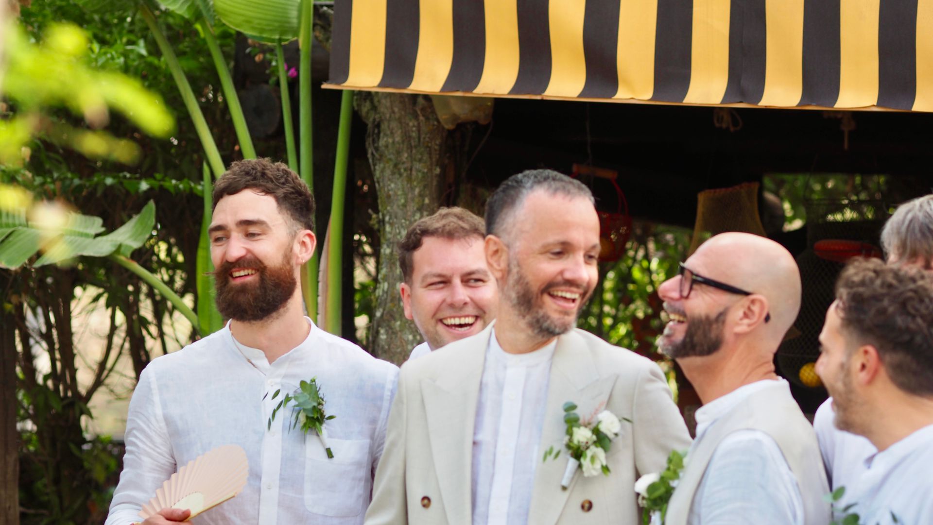 Smiling men in white suits with boutonnieres gathered outdoors under a black-and-yellow awning