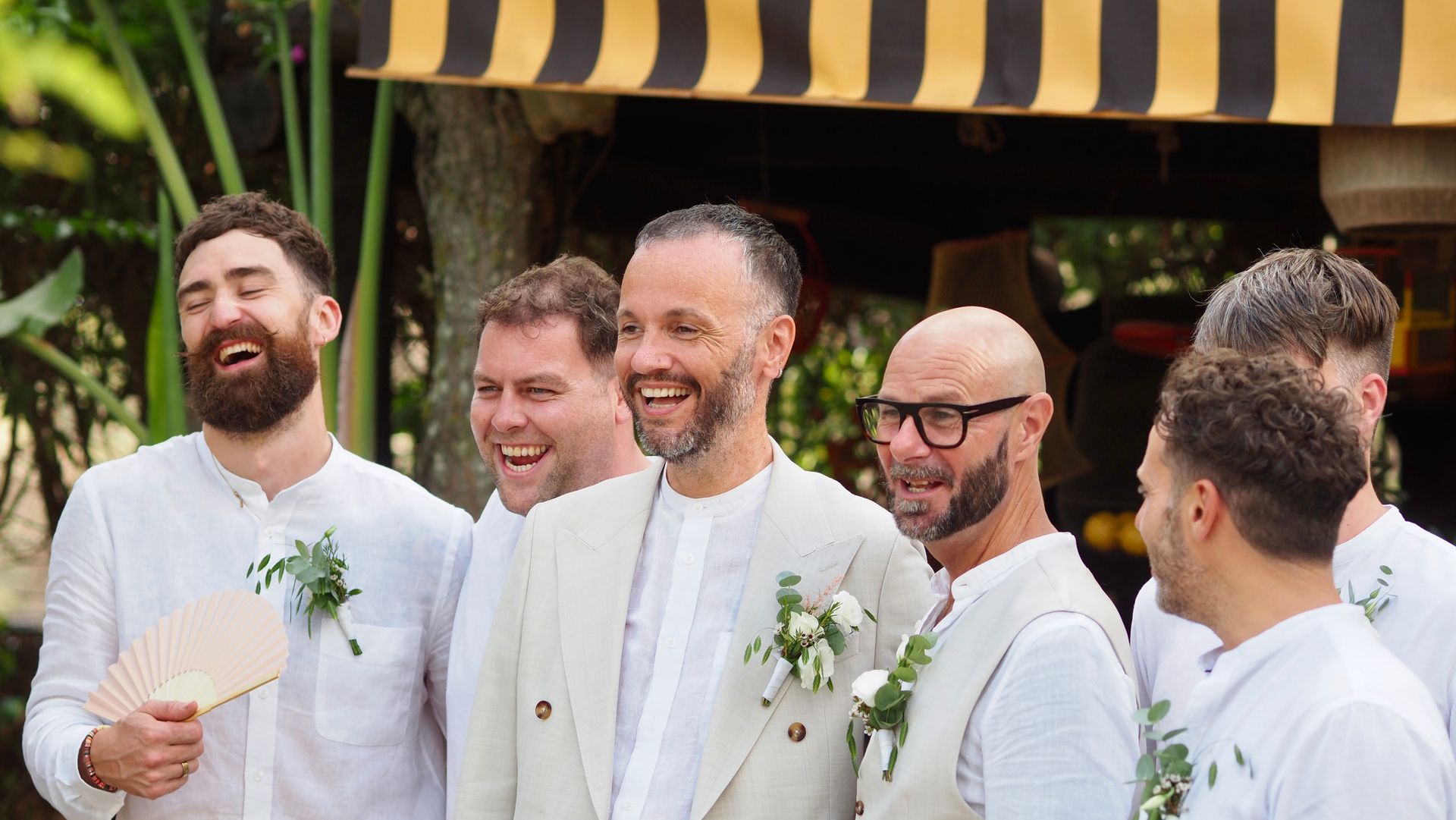 Five people in white jackets and bow ties smiling outdoors under a striped awning, holding drinks.