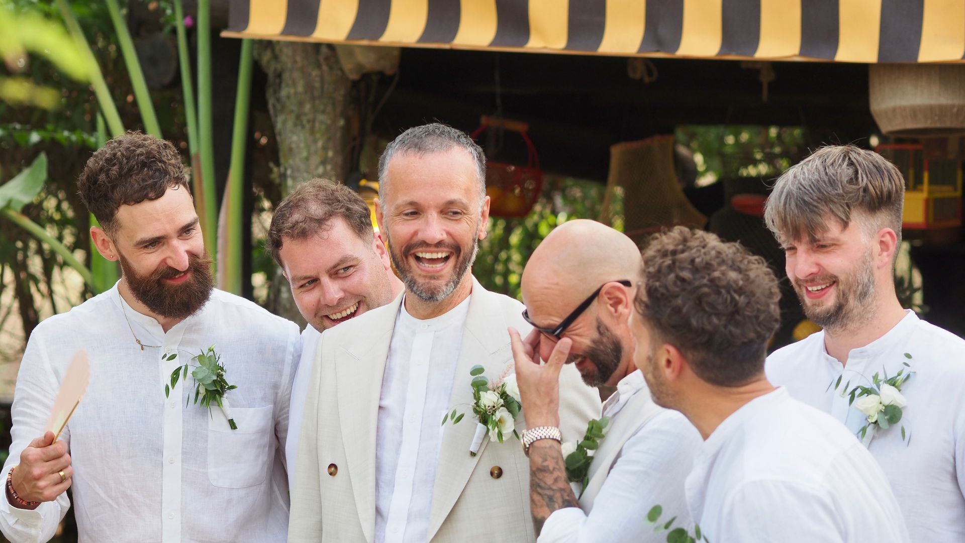 Five smiling people in white suits chatting outdoors under a striped canopy