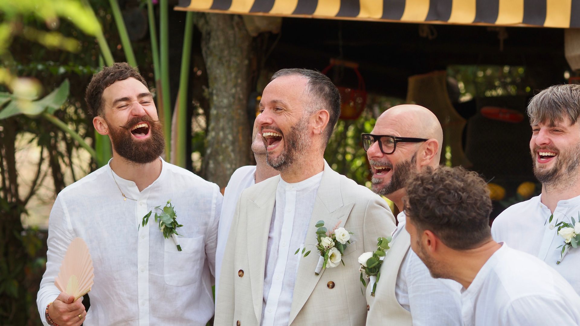 Four men in white suits laughing together at an outdoor celebration under a striped canopy