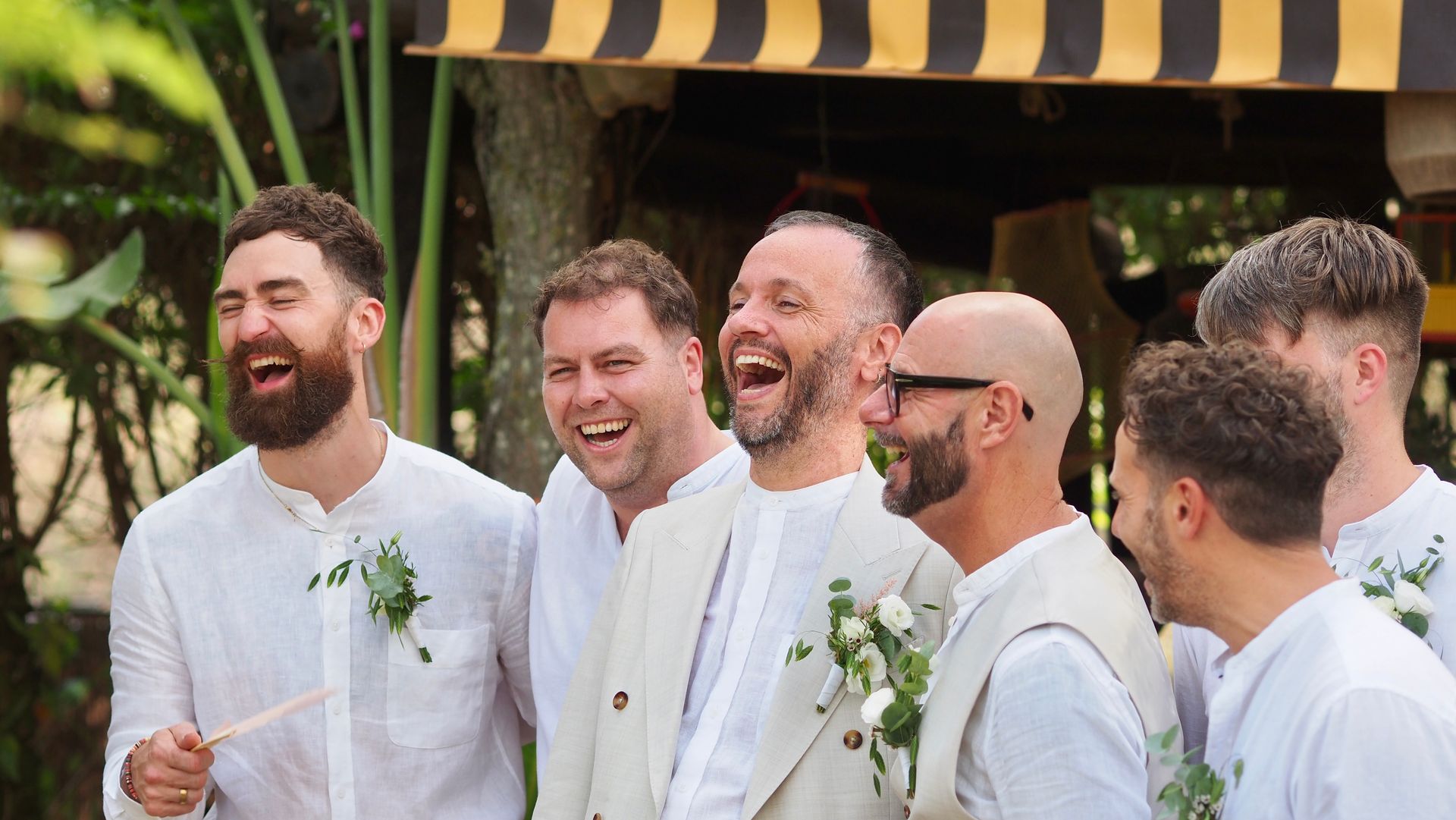 Smiling friends in white outfits standing outdoors at a celebration with striped awning overhead