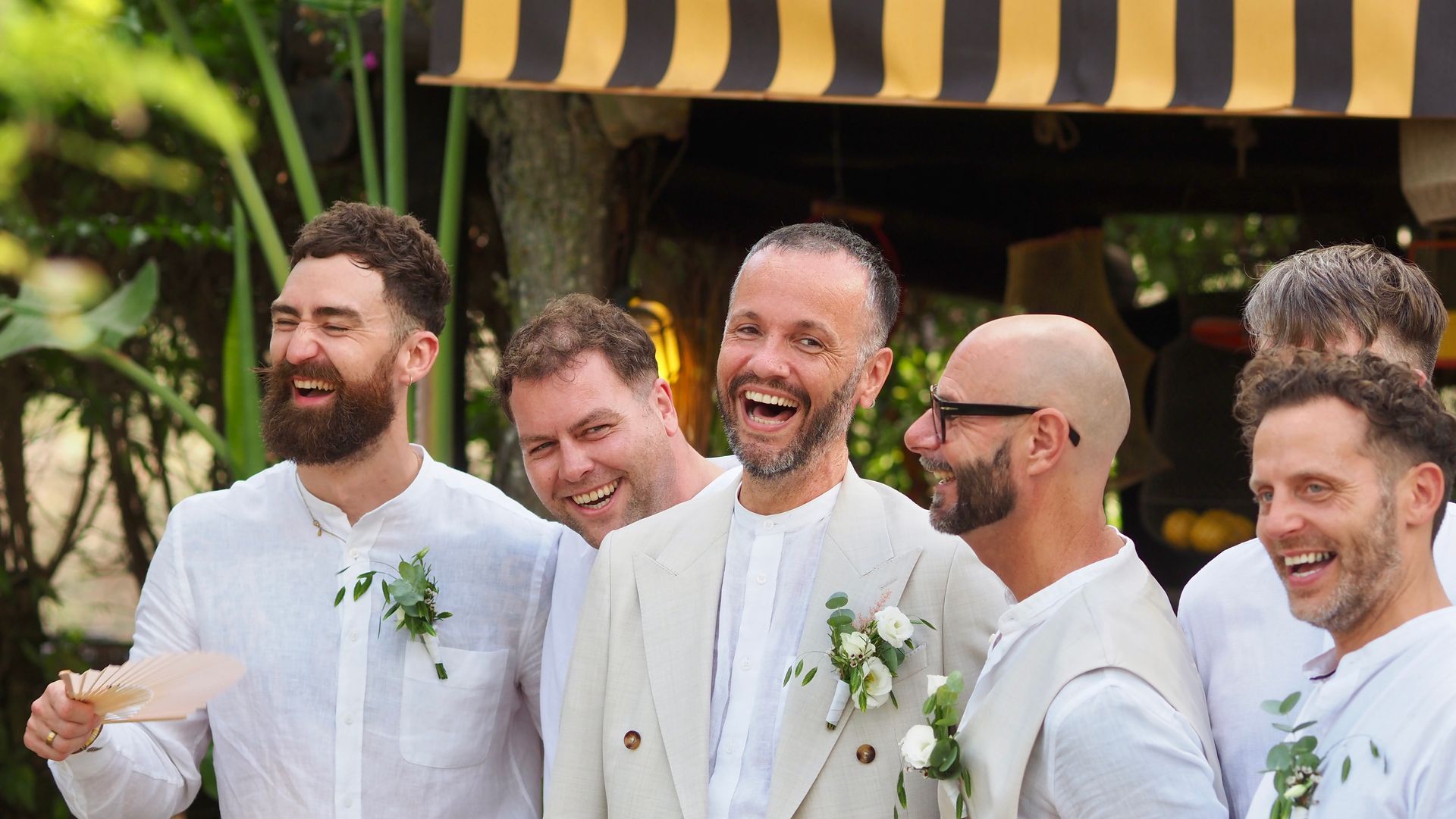 Smiling group in white shirts standing outdoors at a celebration, laughing together near a striped canopy.