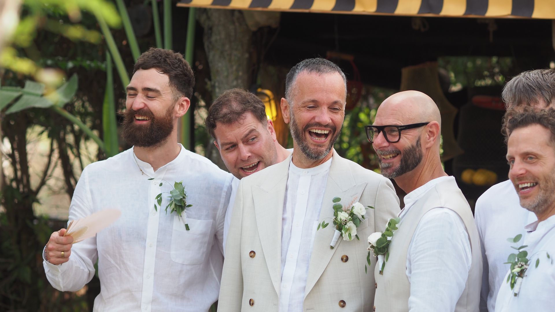 Five men in white suits smiling and laughing at an outdoor celebration.