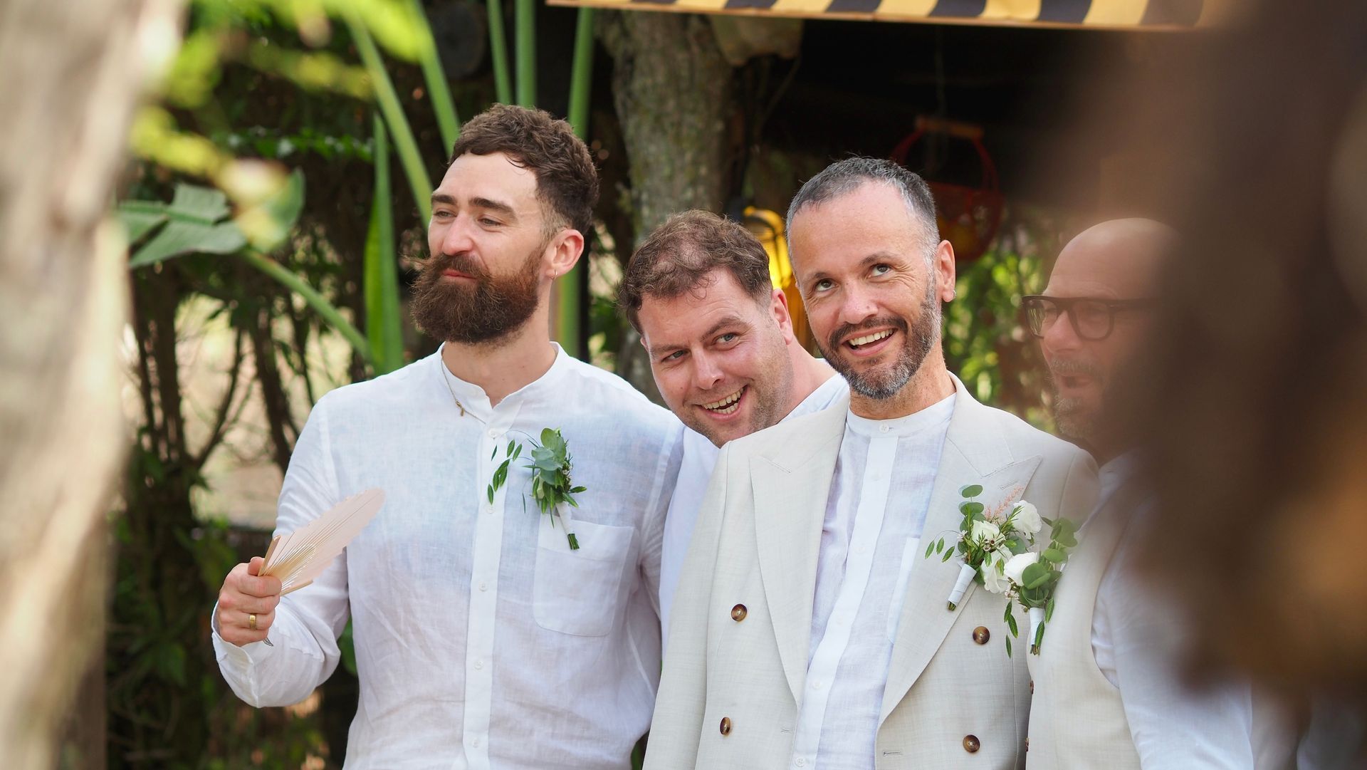 Three men in light suits smiling at an outdoor wedding ceremony