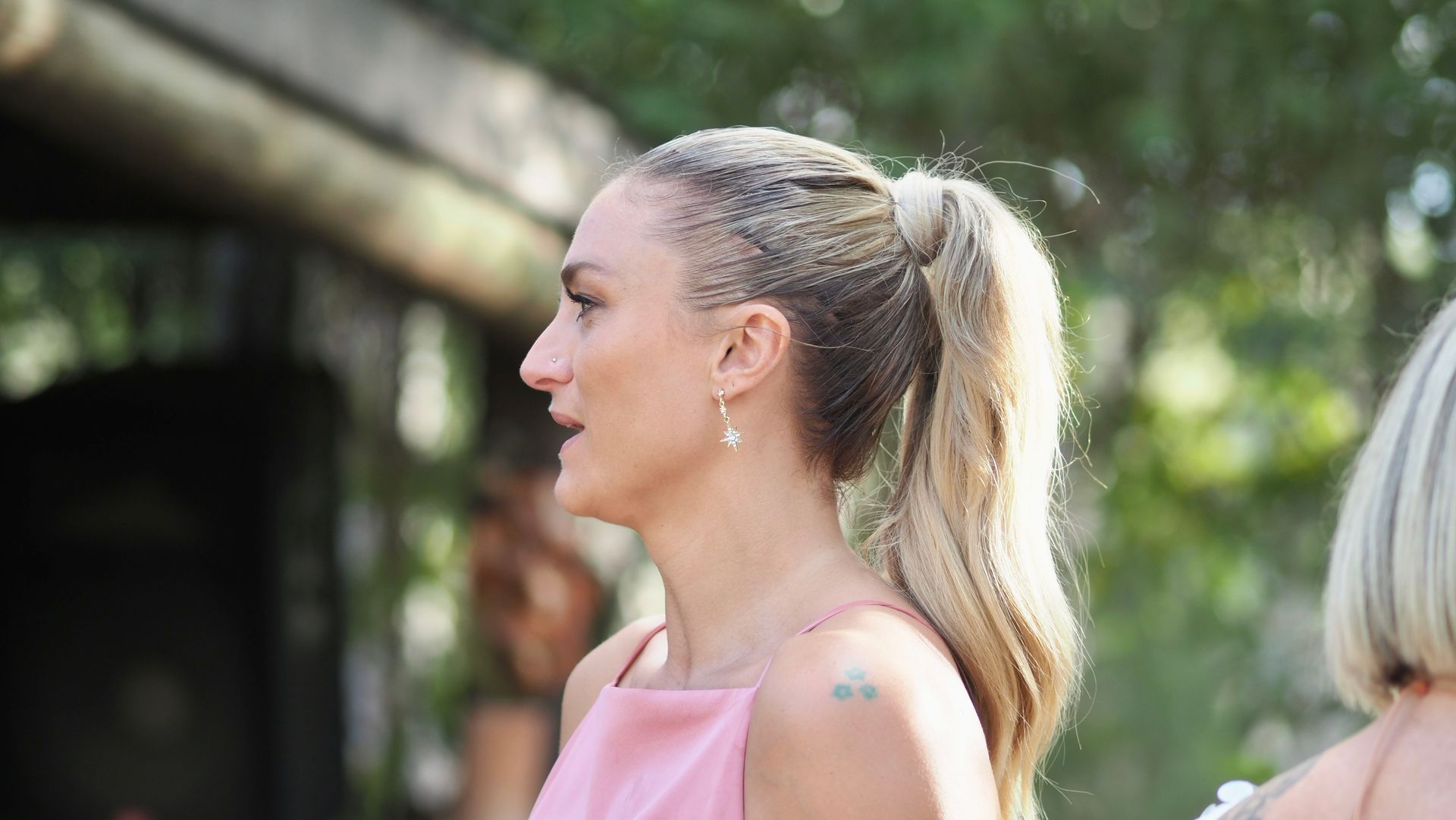 Side-profile of a blonde woman in a pink top outdoors, with trees and another person blurred in the background