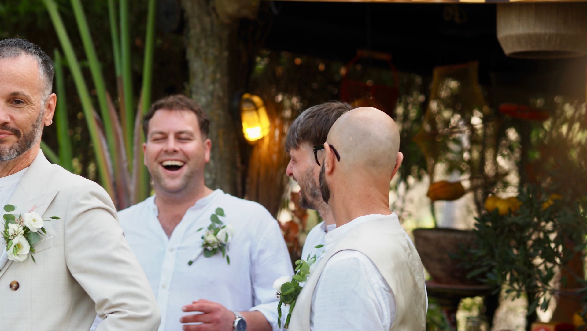 Three men in white shirts and boutonnières laughing together at an outdoor celebration