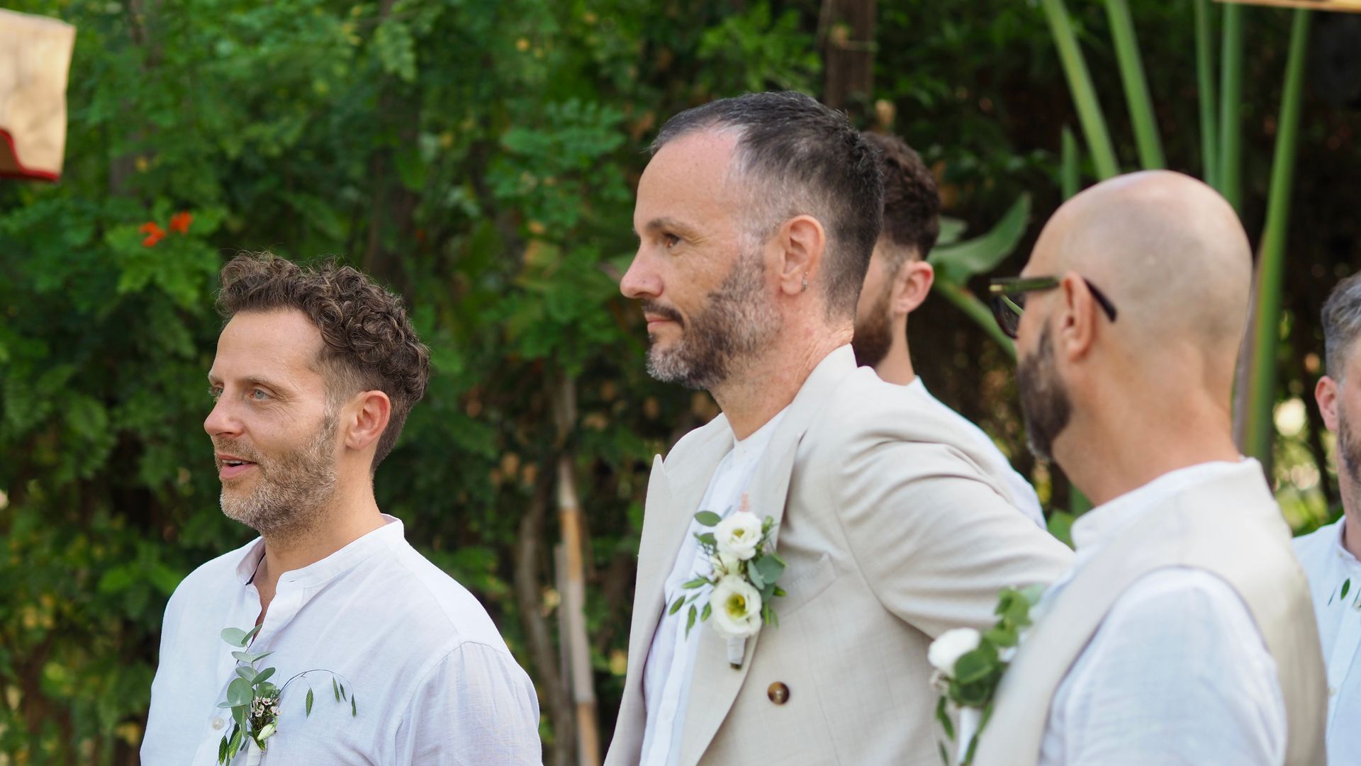 Three men in white suits stand outdoors, smiling in a garden setting.