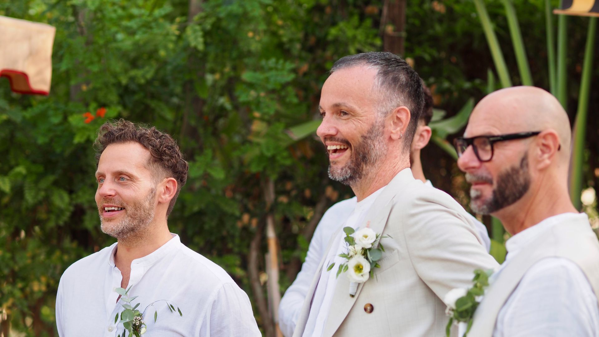 Three smiling men in white jackets stand outdoors at a celebration.