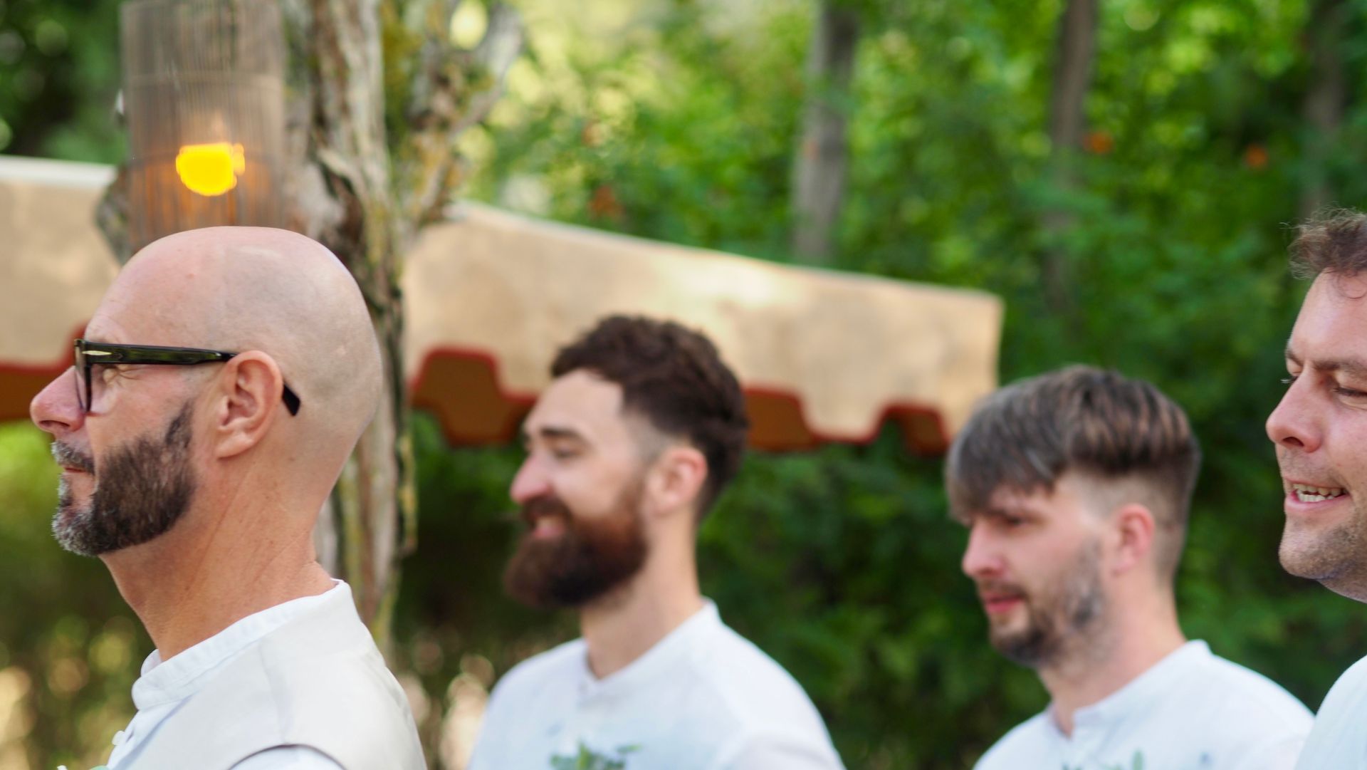 Three men in white shirts stand outdoors with trees and a tent in the background.