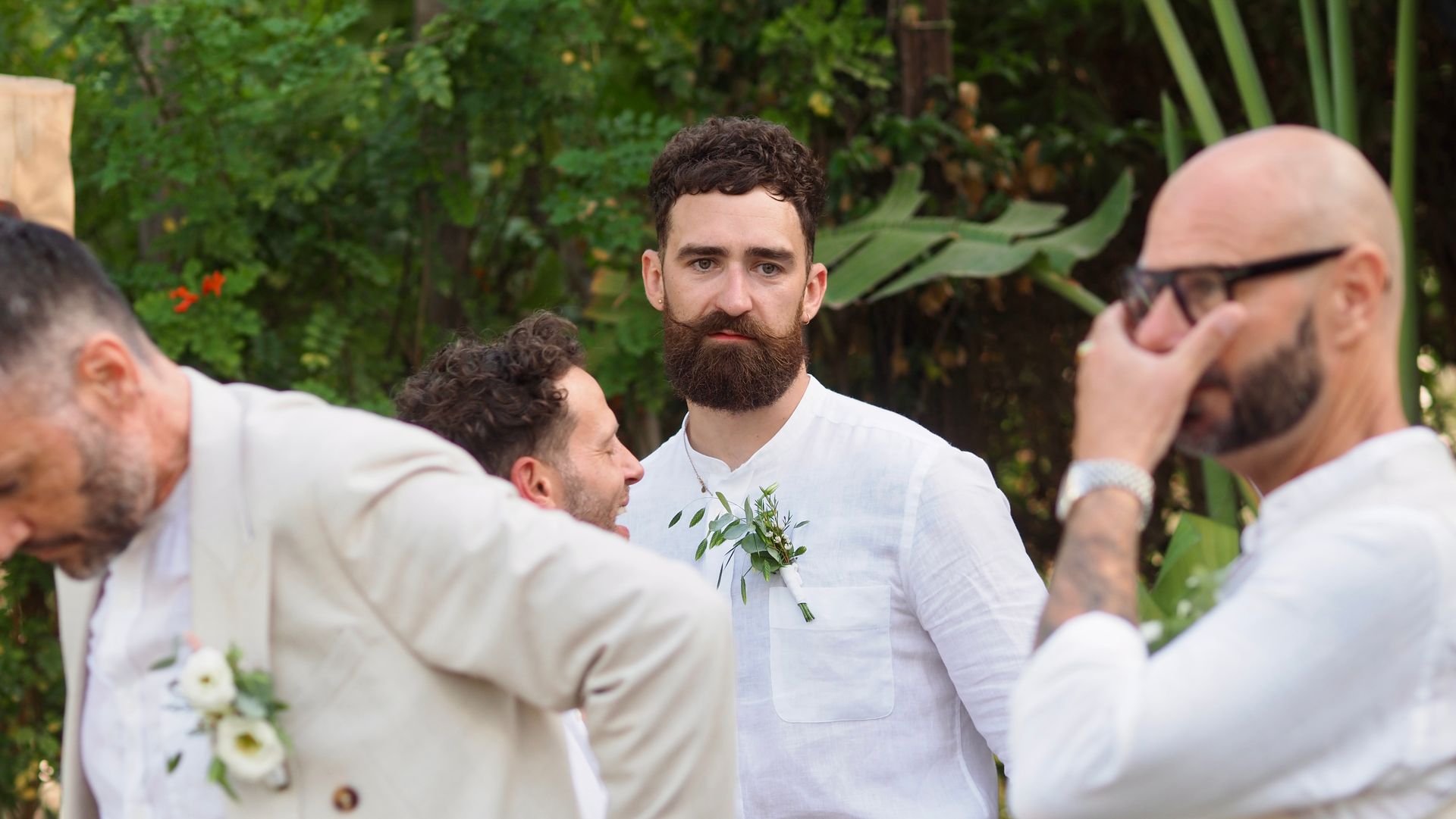 Bearded man in a white shirt stands outdoors among others in light-colored clothing and a bald man with sunglasses.