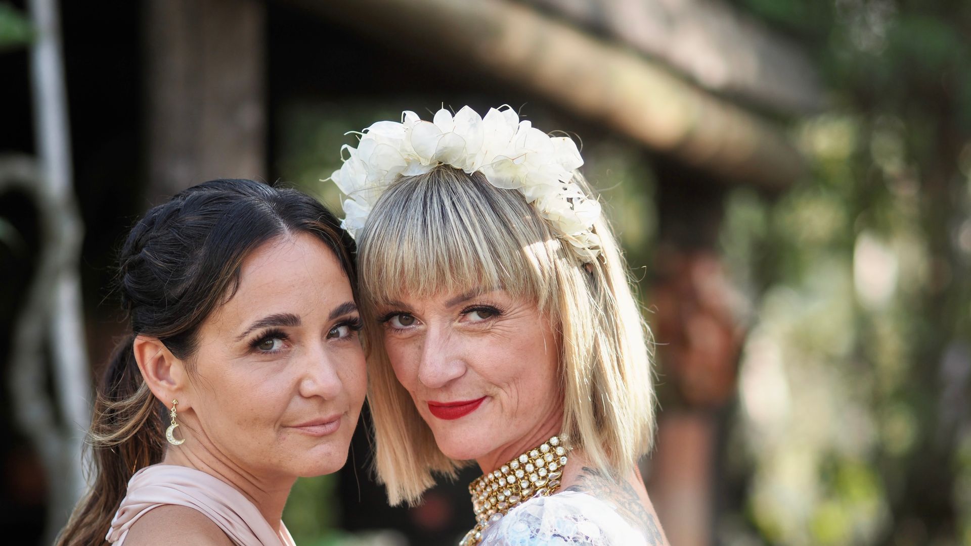 Two women in white attire pose outdoors, one wearing a floral headpiece, both smiling softly.