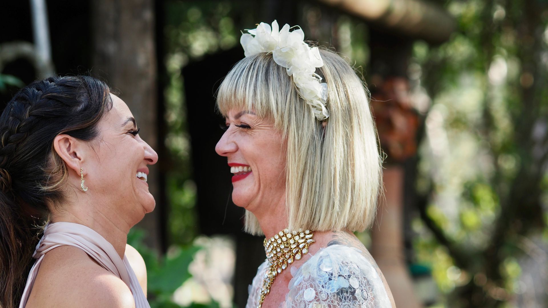 Two women smiling face-to-face outdoors, one in a white lace dress and floral headpiece.