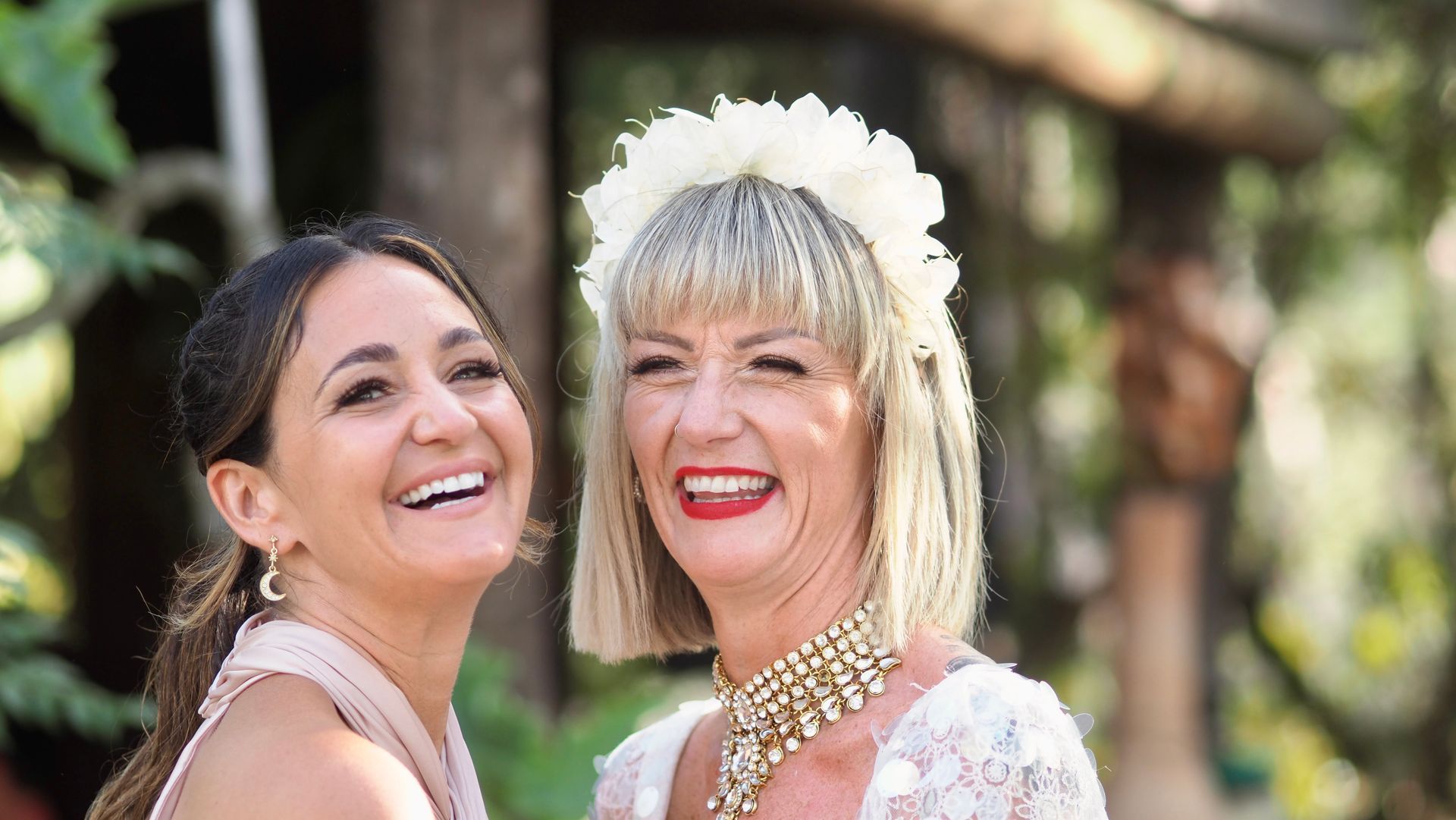 Two smiling women in formal dresses at an outdoor event, one wearing a white floral headpiece.