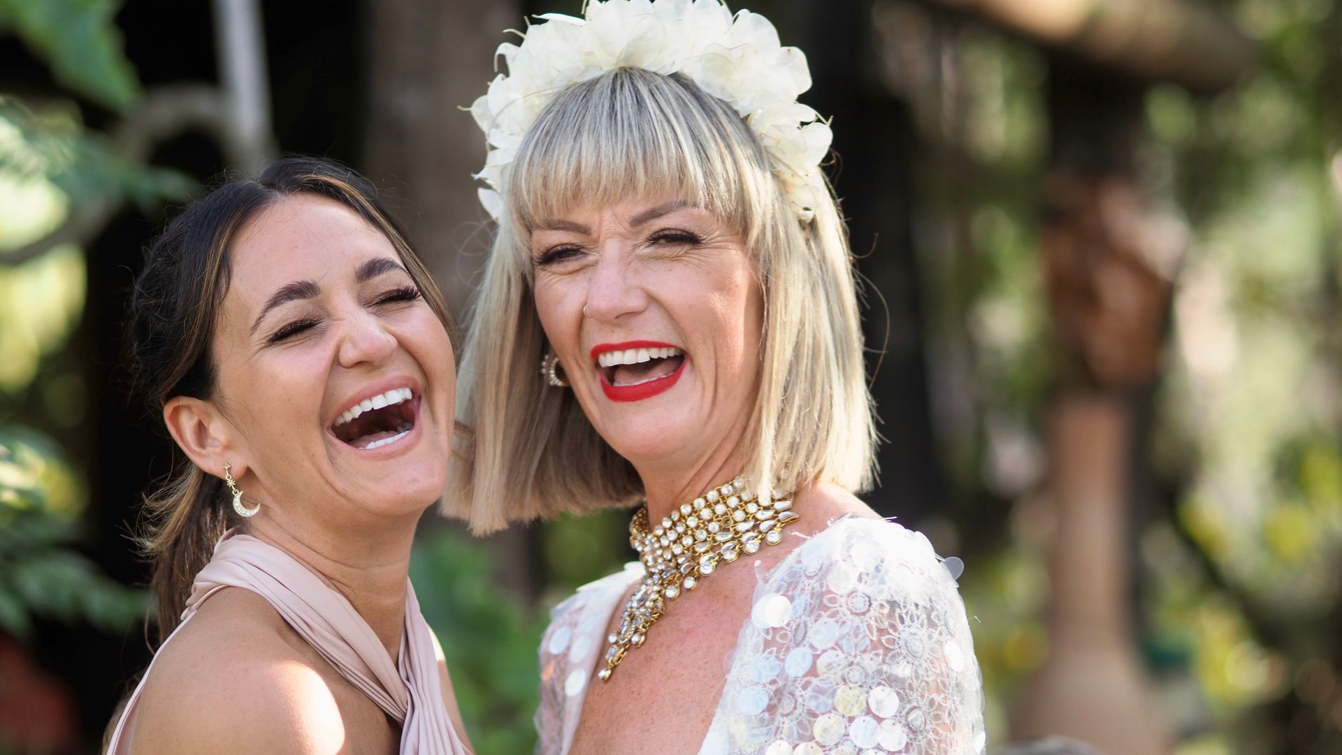 Two women smiling and laughing outdoors, one in a white floral headpiece and dress